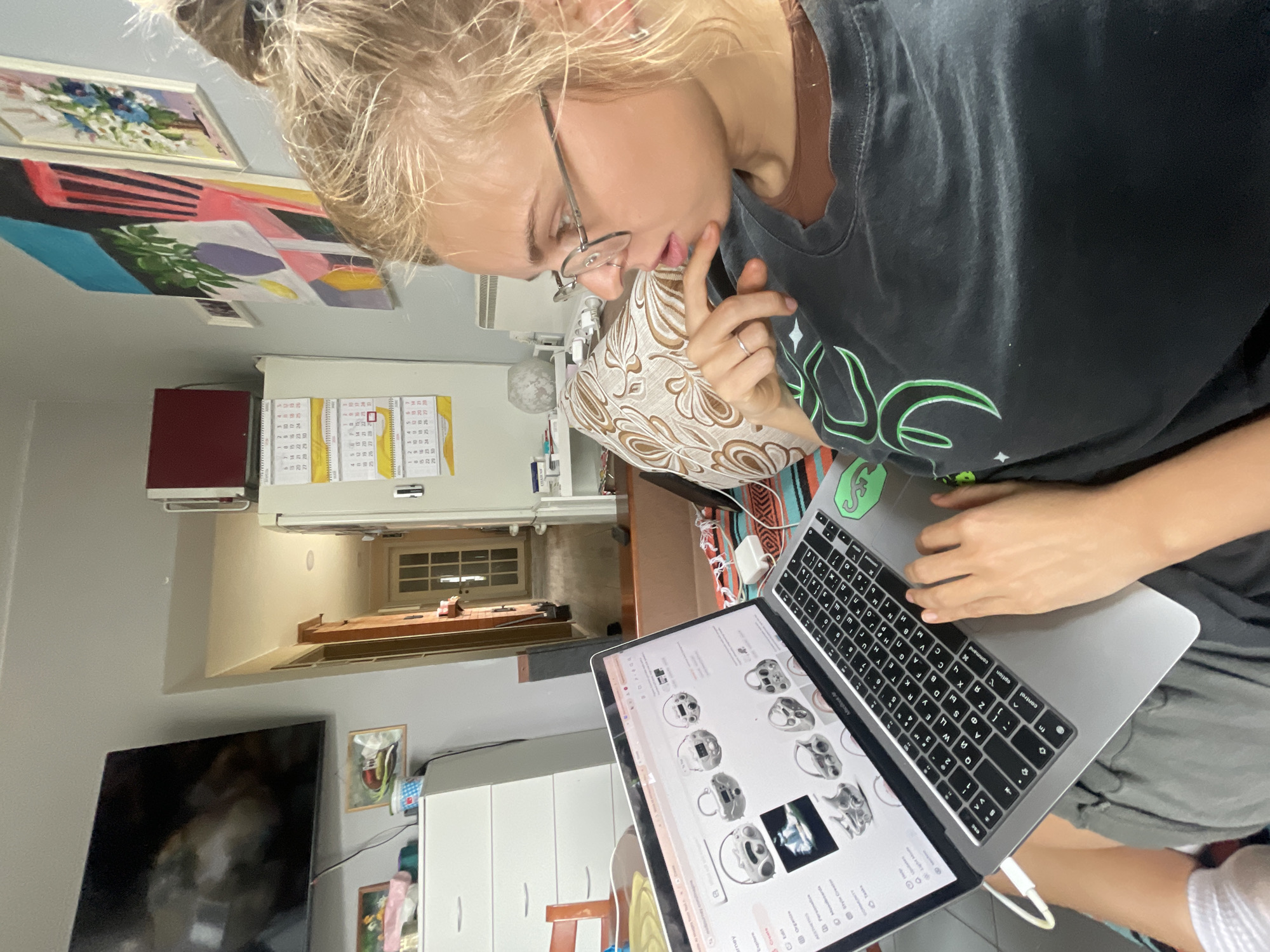 A person with blond hair wearing glasses and a black t-shirt is sitting and using a laptop. The laptop is displaying a website with multiple images of what appear to be decorative items. The person has their left hand near their face and is looking down at the screen. The laptop is resting on the person's lap.
In the background, there is a room with various items, including a television, a white cabinet, and a small table. There are paintings on the wall and a calendar. A doorway leads to another room. It is not possible to determine the location in the city of Sestroretsk, Russia, based on the image.