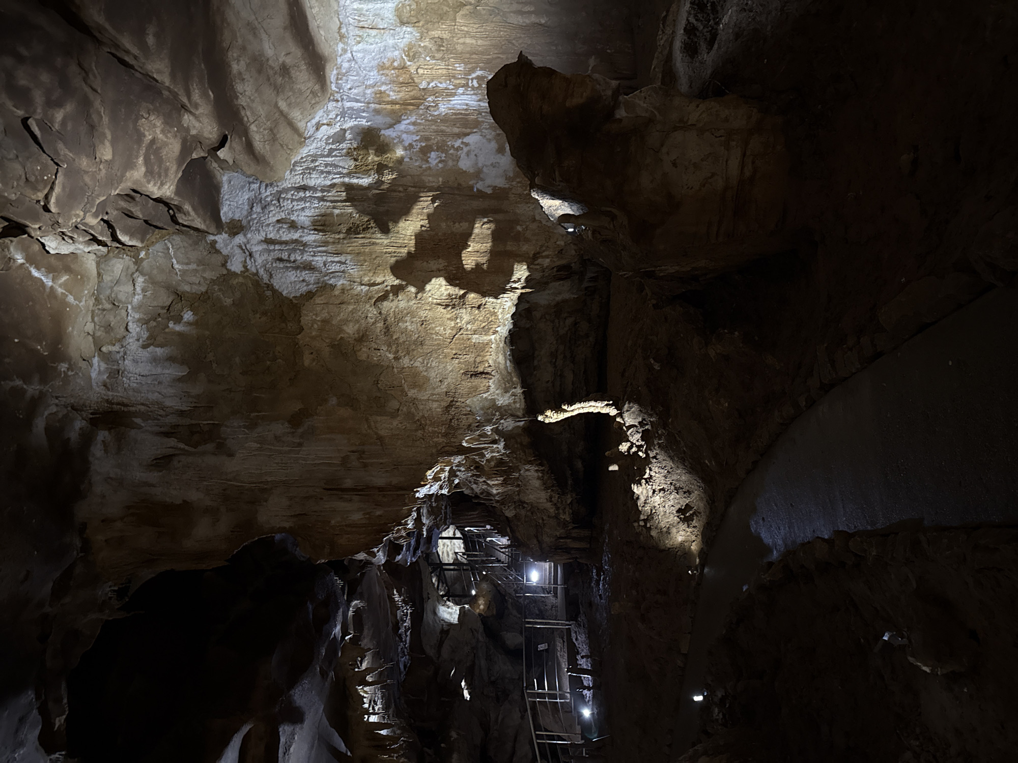 The image shows the interior of Prometheus Cave, located near Tskaltubo, Georgia. The cave's geological formations, consisting of stalactites and stalagmites are illuminated by artificial light. A metal staircase and walkway have been constructed to facilitate visitor access and navigation within the cave. The environment is dark and cavernous, with textured rock surfaces. There are no people visible in the image.