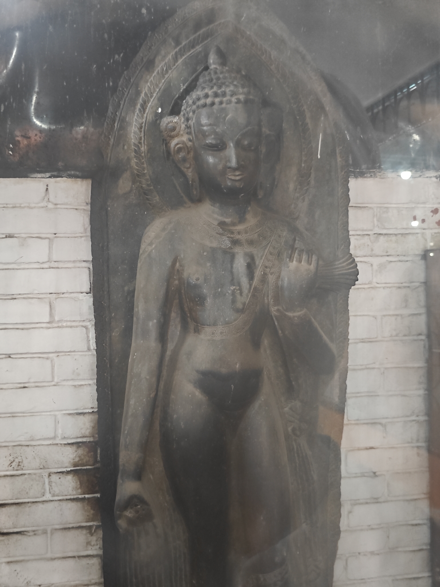 A full-body, dark gray stone statue of a standing figure, likely Buddha or a deity, is encased behind glass. The figure is depicted within a stone niche with carved decorative elements around the head and shoulders. The figure has a serene facial expression, elaborate headdress, and is adorned with necklaces and garments. The statue stands against a background of white brickwork, with some of the brickwork appearing stained or dirty. The scene is dimly lit, with reflections visible on the glass surface, suggesting it is displayed indoors, possibly in a museum or temple within Kathmandu Metropolitan City, Nepal. The overall condition of the statue appears to be aged, with some signs of wear and tear.
