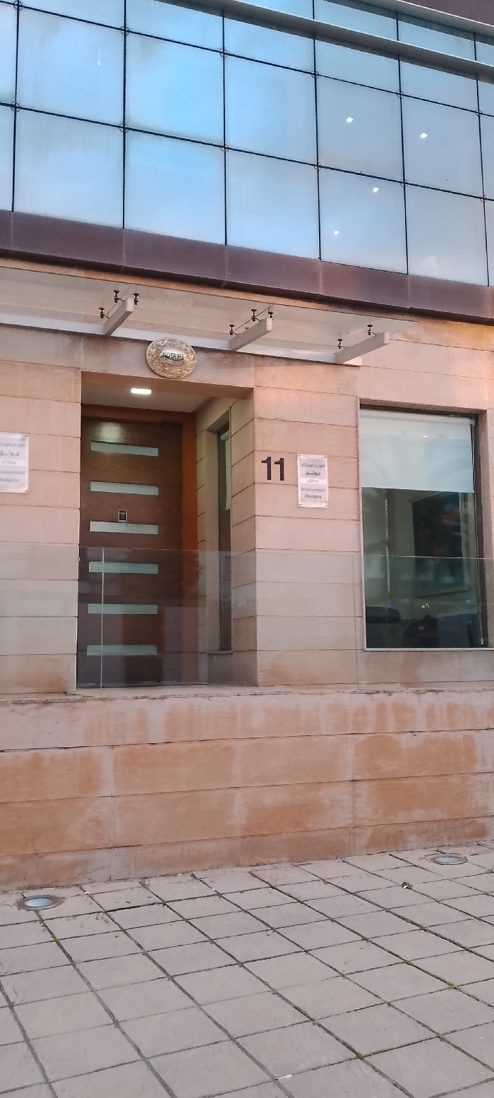 The image displays the ground-level exterior of a modern building, likely an office or commercial establishment, in Aïn El Aouda, Morocco. The building's facade consists of light-colored, horizontally stacked stone cladding on the lower section, with a large grid of blue-tinted glass panels forming the upper part.
The main entrance features a wooden door with multiple horizontal frosted glass inserts. Above the door, a prominent round golden sign reads "NOTAIRE" (French for Notary) along with Arabic script. To the left of the door, a rectangular sign contains Arabic text and "Notaire." To the right, the address number "11" is clearly visible, beneath which another rectangular sign displays Arabic text, likely "مكتب موثق" (Notary Office), and "Notaire." A transparent glass barrier stands in front of the door and an adjacent window, and a glass canopy supported by metal brackets extends over the entrance.
The pavement in front of the building is composed of square light-colored tiles, embedded with circular ground lights. No individuals are present, indicating a quiet scene. The soft, diffused lighting suggests either late afternoon, early morning, or an overcast day. The use of both French and Arabic on the signage aligns with the provided location context of Morocco, confirming the establishment as a notary office.