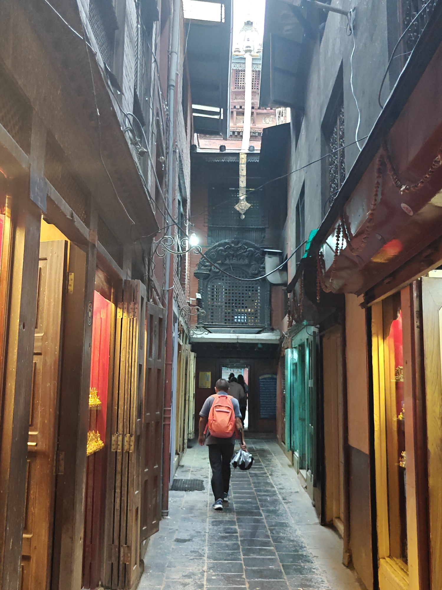 The image shows a narrow alleyway in Lalitpur, Nepal, with tall buildings on either side. A person with an orange backpack is walking away from the camera down the alley. The buildings have wooden doors and windows, some of which are open, revealing red curtains and ornate carvings. The alleyway is paved with stone tiles and there are some potted plants visible. In the background, a tall pagoda-style building with intricate architecture can be seen. The lighting suggests it is daytime, possibly late afternoon, with some areas in shadow and others illuminated by sunlight. The overall impression is of a bustling, ancient city with a rich cultural heritage.