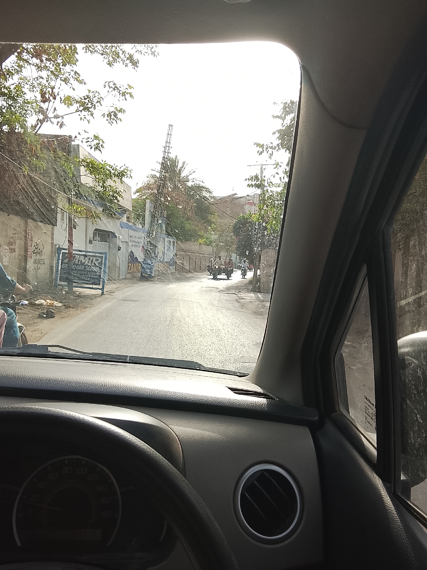 Captured from inside a vehicle, the image shows a street scene in Gulgasht Colony, Pakistan. The road is paved and appears sunlit. On the left side of the road is a building with a sign that reads "ZAMIR GRAMMAR SCHOOL." A person on a motorbike is visible near the building. Further down the road, two motorbikes are driving, and some pedestrians are on the sidewalks. Overhead utility lines are strung between poles. The sky is bright, suggesting daytime. The foreground shows the dashboard of the car, including the steering wheel, speedometer, and air vent.