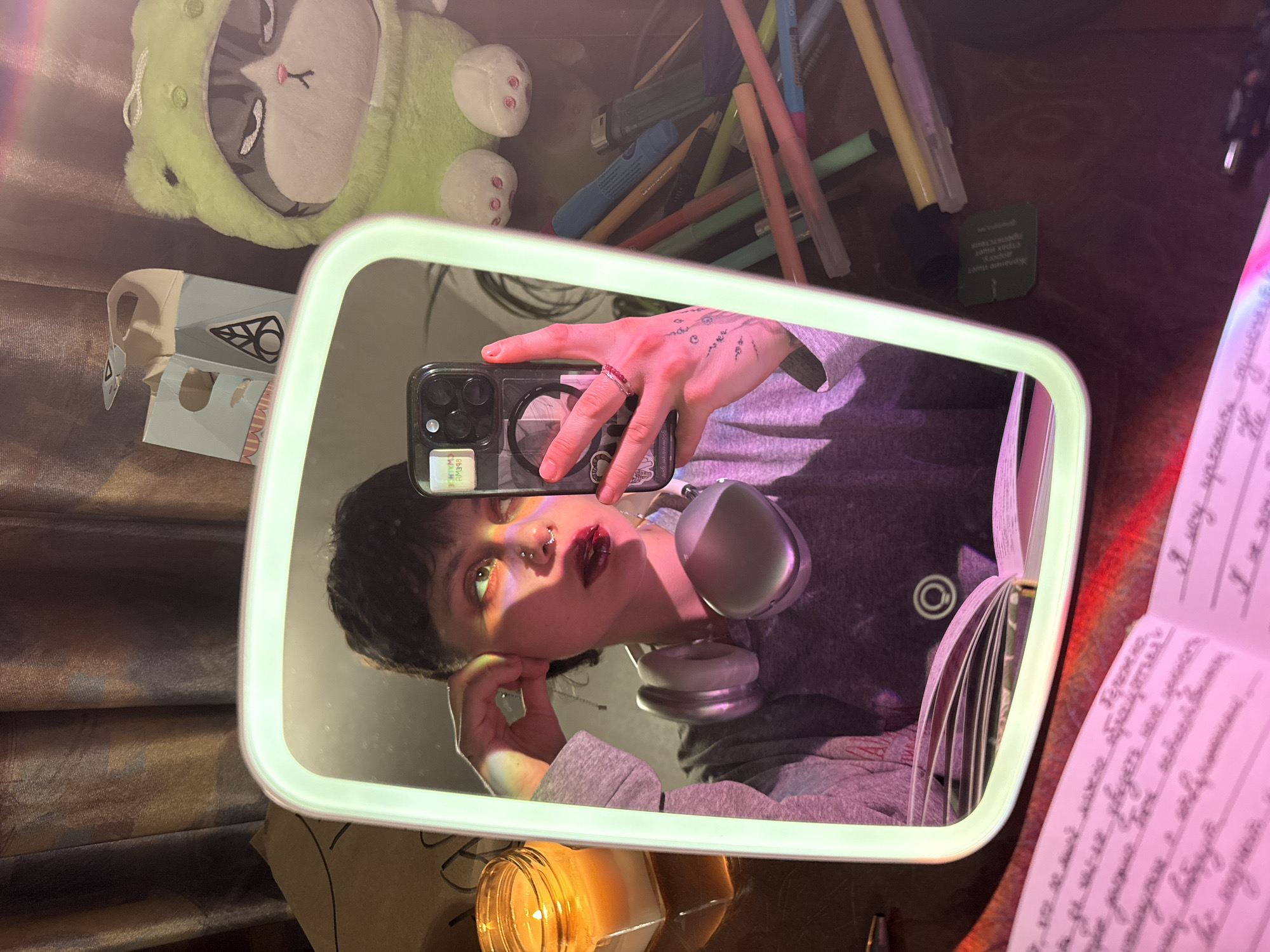 Here's a detailed description of the image:
**Scene Overview:** The image captures a person taking a selfie in a lighted mirror. The environment appears to be indoors, possibly a bedroom or study area, given the presence of personal items.
**People:**
* The primary subject is a person reflected in the mirror. The person has dark hair, wears silver headphones around their neck, and has facial piercings (nose). They are wearing a gray hoodie. Their hand is visible holding a smartphone.
**Objects:**
* **Mirror:** A rectangular mirror with an integrated LED light frame is the central object.
* **Smartphone:** The person is holding a smartphone to take the selfie.
* **Headphones:** Silver over-ear headphones rest around the person's neck.
* **Notebook:** An open notebook with writing on the pages is visible on the table, to the right of the mirror. The text appears to be in Russian.
* **Candle:** A lit candle in a jar is situated at the bottom of the mirror.
* **Writing Utensils:** A collection of colored markers and pens are arranged in the upper right corner.
* **Stuffed Animal:** A green plush toy is partially visible in the upper left.
* **Box:** A small box with printed designs sits near the plush toy.
* **Misc:** There are some other small objects that are unidentifiable.
**Actions and Interactions:**
* The person is taking a selfie using their smartphone.
* The person is holding the phone up to the mirror, with their hand and part of their arm visible in the reflection.
**Environment:**
* The scene appears to be lit with a mix of natural light and the light from the mirror. There also seems to be a purple hue light.
* The surface where the items are arranged seems to be a wooden desk or table.
* A piece of fabric, likely a curtain, is visible in the upper-left corner.
**Location:** There is written Russian text on the notebook in the frame and a card that says "Made in Saint Petersburg, Russia." Therefore, the location may be the city of Saint Petersburg, Russia.