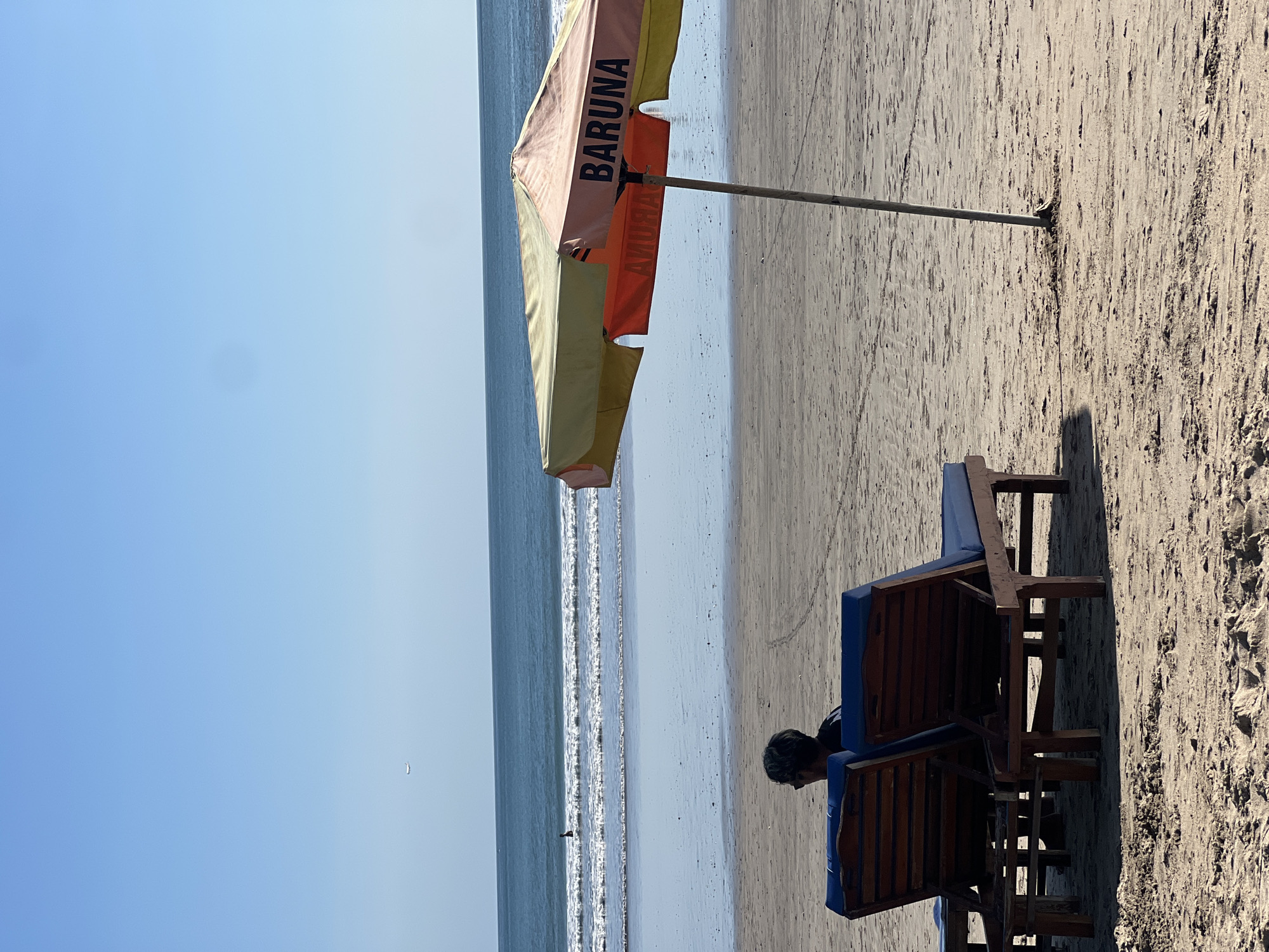 A person sits on a wooden beach chair with a blue cushion, looking down towards the sand. The chair is positioned on a sandy beach with the ocean in the background. The water is calm with small waves breaking near the shore. The sky is clear and blue, suggesting a sunny day, likely in the late morning or early afternoon given the angle of the shadow cast by the umbrella.
An open beach umbrella provides shade. The umbrella has alternating panels of light beige and orange, with black text printed on it. The text "BARUNA" is clearly visible in black letters on one of the light beige panels. Another word, "AMURAB," is visible in reverse on an orange panel, suggesting it may be on the underside of the umbrella and facing the photographer, or a reflection.
In the distance, a small figure can be seen in the water, appearing to be standing or wading. The overall scene evokes a sense of relaxation and a typical beach day in Kuta, Indonesia. The lighting and shadows indicate it is daytime.