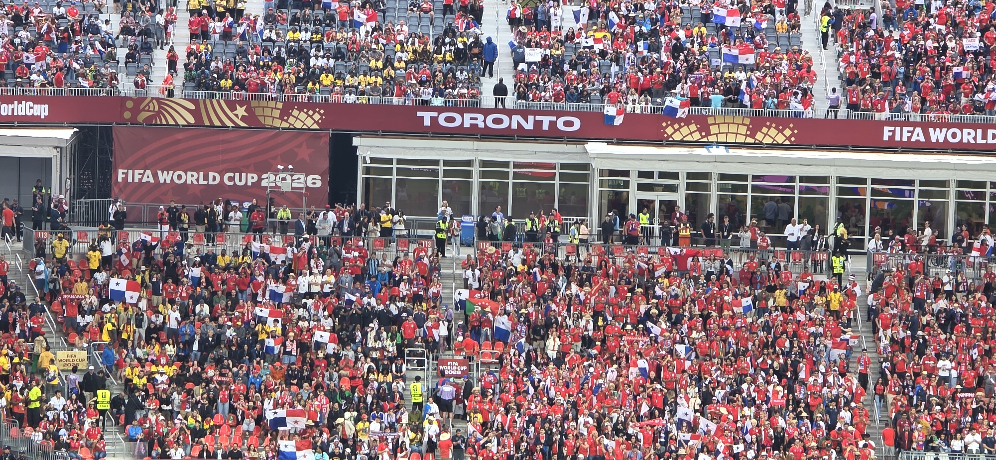 This image depicts a crowded outdoor stadium filled with spectators, predominantly wearing red, cheering and waving flags. A large banner at the top of the stadium reads "FIFA WORLD CUP 2026 TORONTO". The architecture suggests a modern sports venue with tiered seating, covered areas, and visible support structures. Security personnel in high-visibility vests are present among the crowd, managing the flow of people and ensuring safety. The atmosphere appears festive and energetic, indicative of a major sporting event. The presence of the FIFA World Cup 2026 branding and the city name "TORONTO" clearly identifies the location as a stadium in Toronto, Canada, preparing for or hosting a FIFA World Cup 2026 event. The flags are predominantly red and white, with some featuring stars and blue accents, possibly representing national teams or supporter groups. People are standing, seated, and some are captured mid-action, indicating active engagement with the event.