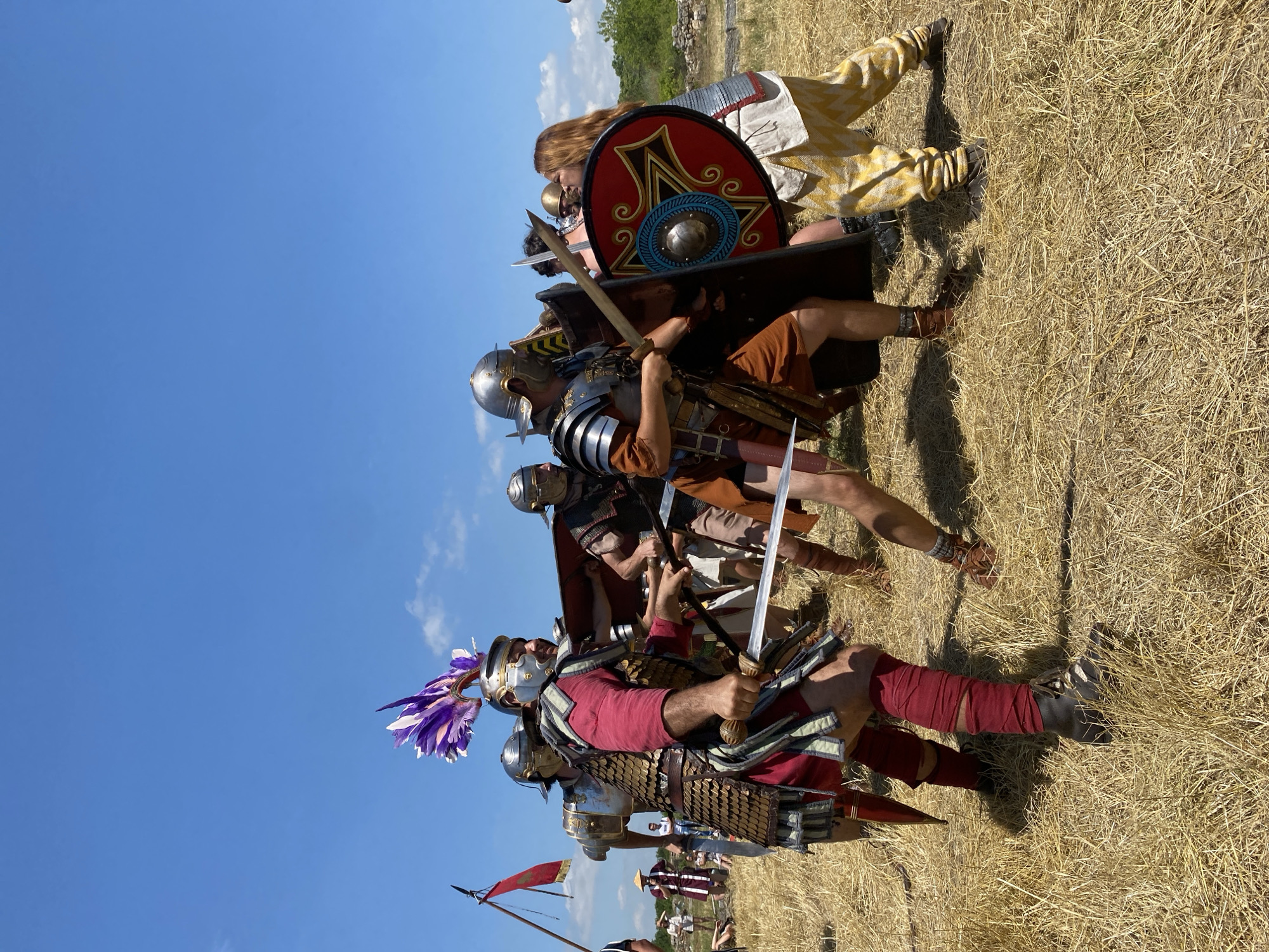 The image captures a historical re-enactment of an ancient combat scene, set in a dry, open field near the ancient Roman city of Oescus, in the vicinity of Gigen, Bulgaria, under a clear blue sky with scattered white clouds.
In the foreground and midground, approximately seven individuals are engaged in a simulated battle. On the left, a participant styled as a Roman legionary wears a red tunic, segmented metallic and possibly leather lamellar armor, and a silver-colored helmet adorned with a prominent purple and white plume. He holds a gladius-style sword in his right hand, pointed towards the right, and is positioned in a dynamic stance with red leg wraps and dark boots visible. Behind him, another Roman-styled participant in lorica segmentata armor and a helmet holds a large rectangular scutum, partially obscuring a red rectangular vexillum on a pole positioned to the far left.
Towards the center-right, a third Roman-styled participant in lorica segmentata and a helmet holds a gladius and a rectangular shield, facing right. He engages with a fourth participant on the immediate right, who has long reddish-brown hair and wears a chainmail-like tunic, light-colored long-sleeved clothing, and distinct yellow trousers with a repeating pattern. This individual holds a round red clipeus shield featuring a complex geometric design in black, blue, and gold tones, centrally punctuated by a metal boss. Behind these two, other figures are partially visible, also in historical attire with shields and swords, contributing to the simulated combat.
The ground is covered with golden-yellow dry grass or straw. In the distant background, several figures are observing the scene, one on the far left wearing a wide-brimmed hat and tunic. Beyond them, low green vegetation and what appear to be remnants of ancient stone structures are visible under the bright sunlight. The overall scene depicts an active, staged confrontation between historical re-enactors.