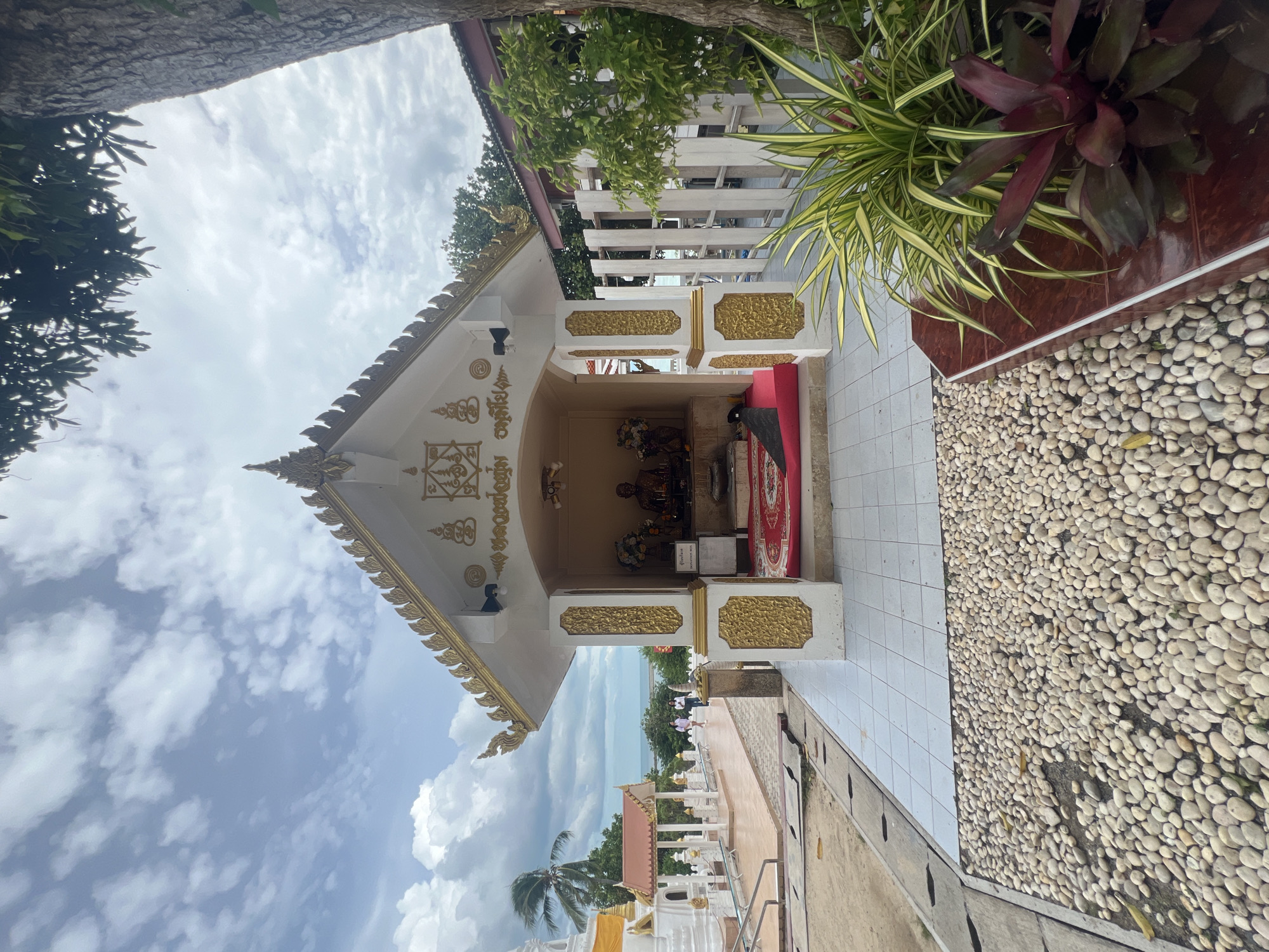 A small, ornate temple structure stands center-frame, painted white with gold trim and decorative detailing. Two seated Buddha statues are visible inside the open shrine, placed on a red rug. To the left, a white stupa with gold accents is partially visible, and in the distance, a body of water can be seen. Two women are walking in the middle ground towards the stupa. The foreground is covered in light-colored pebbles, with a cluster of plants in a planter to the right. The sky is cloudy, with patches of blue.