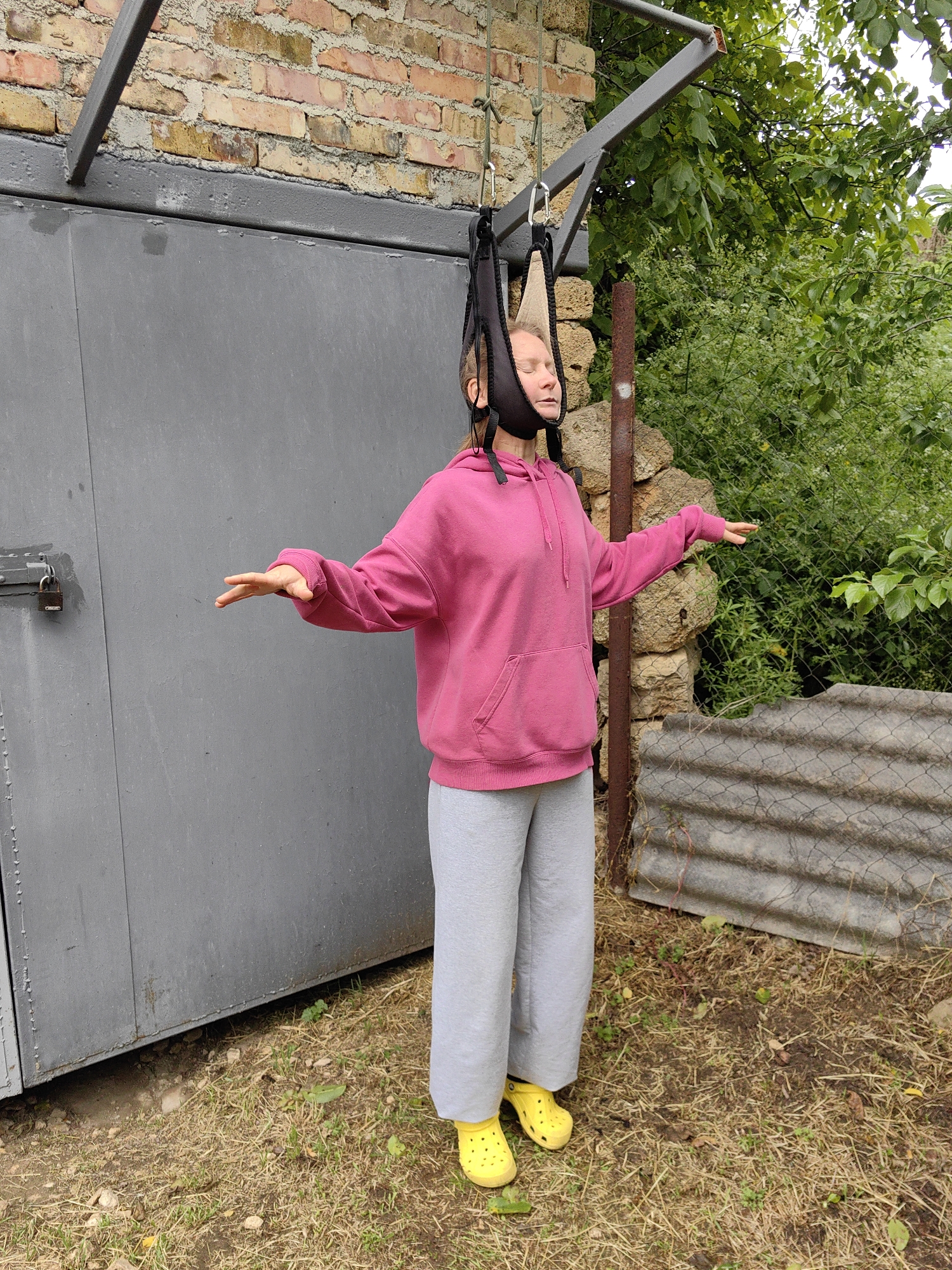 A woman is suspended by her head from a metal frame in Зуйское сельское поселение, Ukraine. She wears a pink hooded sweatshirt, gray sweatpants, and yellow Crocs. Her eyes are closed, and her arms are outstretched. She is suspended from a metal frame attached to a brick and gray metal building. Behind her are shrubs and a metal fence. The ground is covered in dry grass.