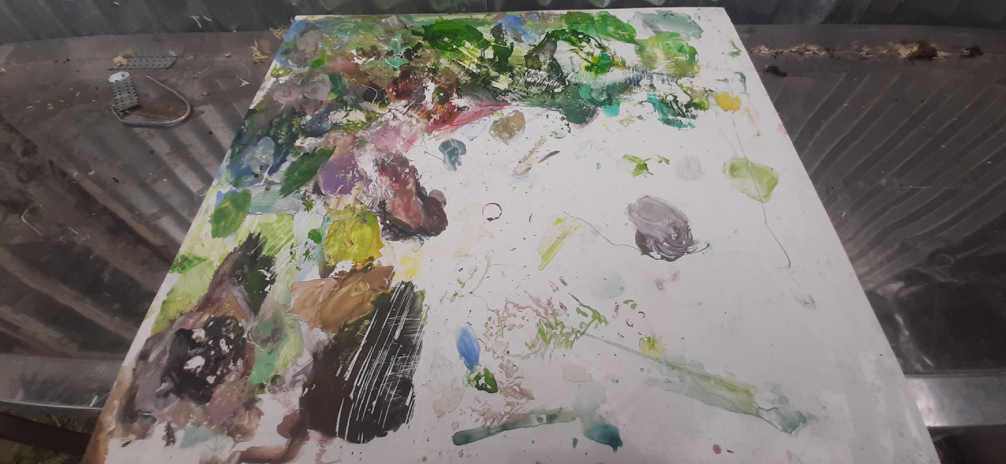 The image shows a white palette covered in a variety of colorful paint splatters and smears. The palette is positioned on a textured, dark surface that appears to be a wooden table or workbench. In the background, a corrugated metal surface is visible. The scene is illuminated by overhead lighting, creating shadows and highlighting the textures of the paint and the surfaces. There are no people or animals present in the image. The location of Yessentukskaya, Russia is not discernible from the provided image.