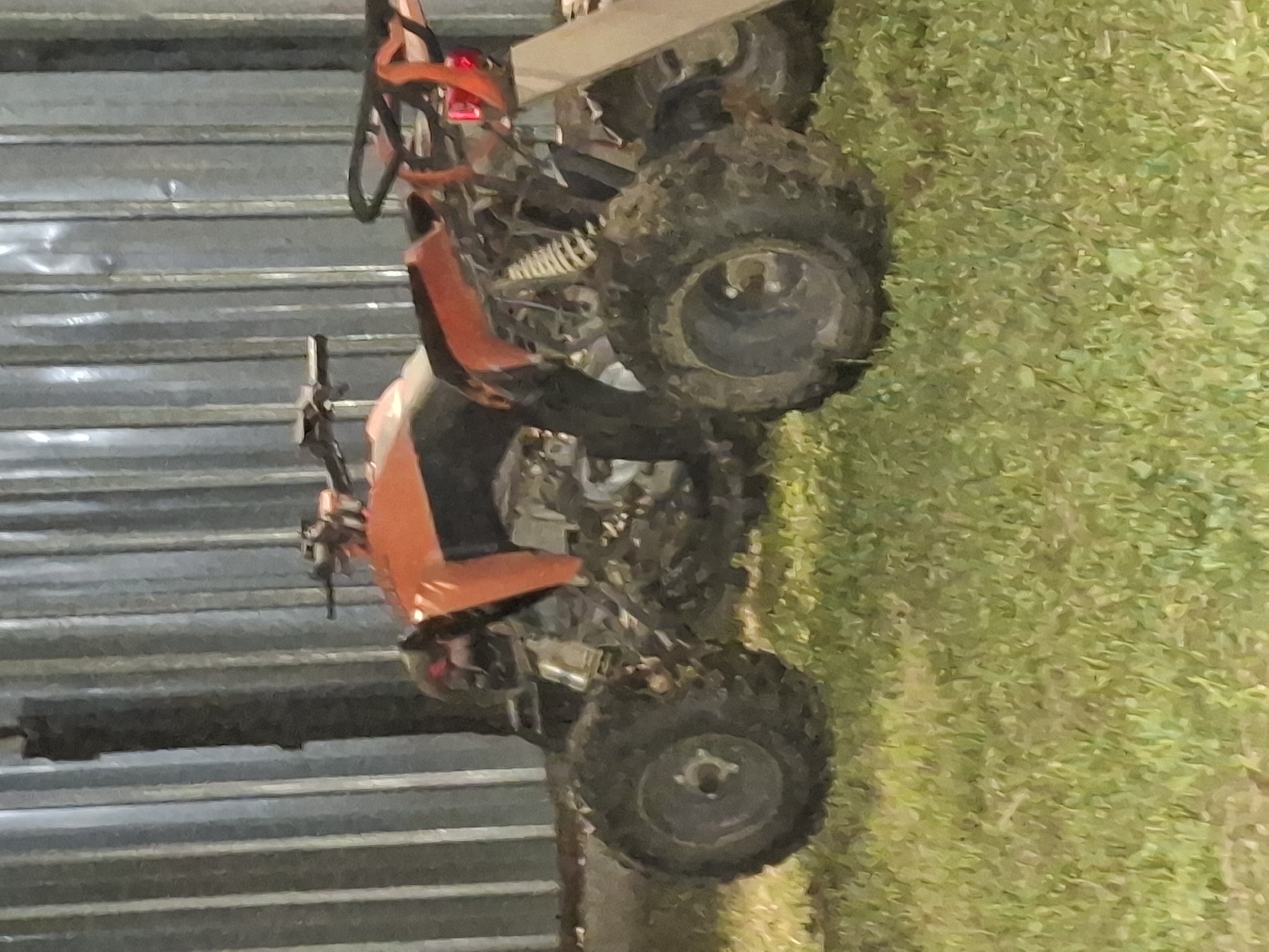 This is a medium shot, low-angle, outdoor photograph of an orange ATV parked on a grassy area in front of a corrugated metal wall. The ATV is positioned diagonally, with its front facing the right side of the frame and its rear towards the left. The ATV appears to be weathered and possibly used, with visible suspension components and thick, knobby tires. The corrugated metal wall, which takes up the background, is vertical and grey. The grass in the foreground is green and appears somewhat dry in places. A dark, vertical post is visible to the left of the ATV, partially obscuring the metal wall. There are no people or animals visible in the frame. The image quality is somewhat grainy and appears to have been taken in low light conditions. The location is specified as Almaty, Kazakhstan.