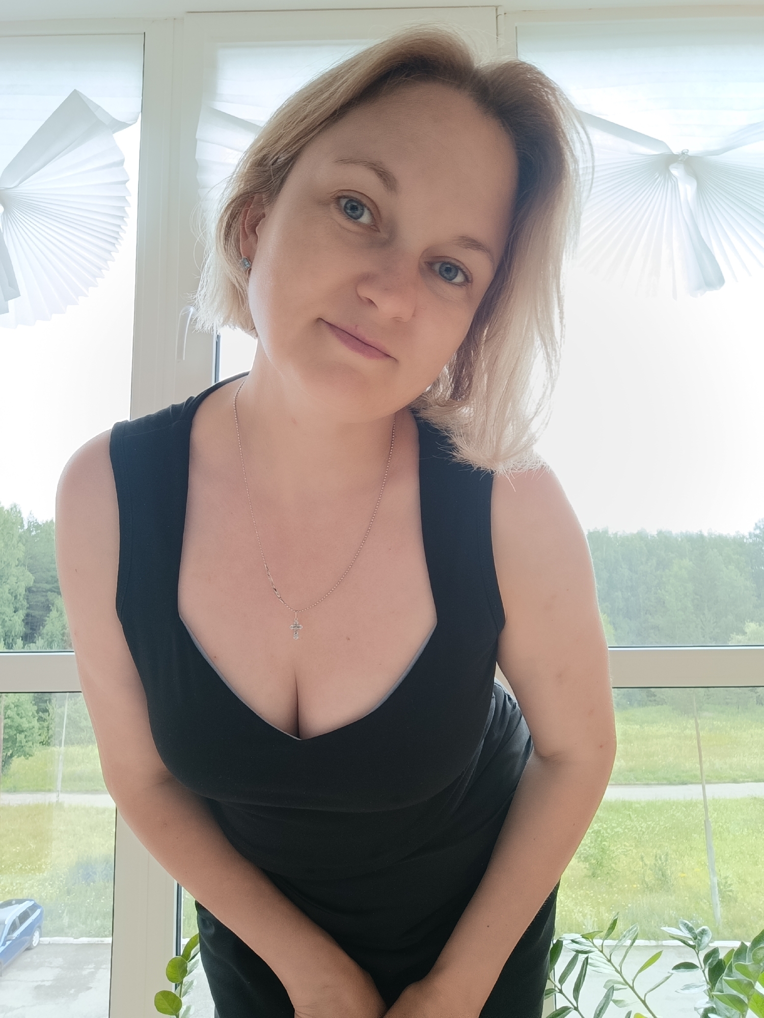 A woman with short, blonde hair and blue eyes is posing for a photo indoors, near a window. She is wearing a black dress with a low neckline and a silver necklace with a cross pendant. Her right hand is partially visible, resting near her chest. Through the window, a green, tree-filled landscape is visible, with a blue car parked in the distance. The window is covered with white pleated blinds. The overall lighting suggests it is daytime. The location is identified as Марковский, Russia.