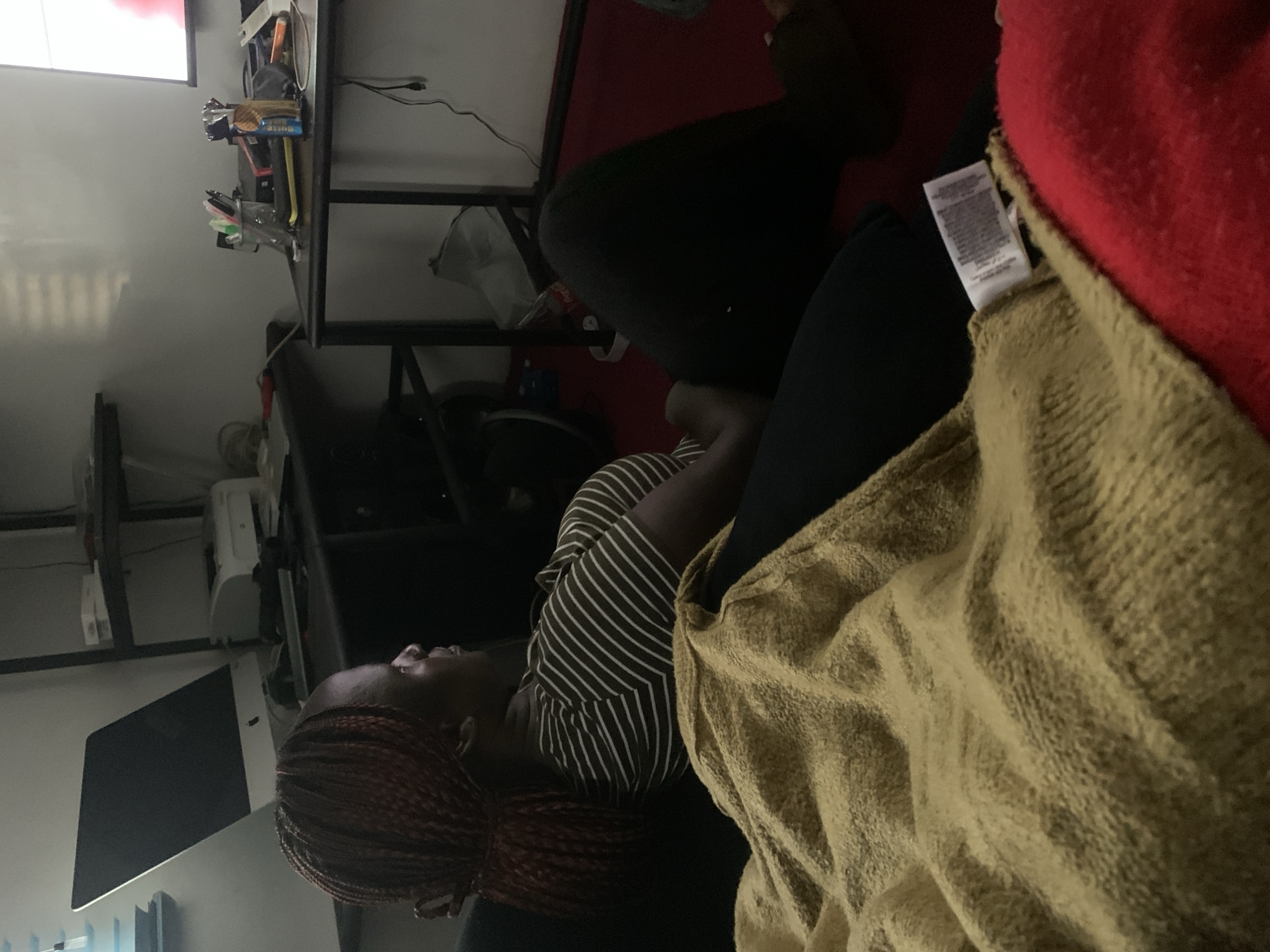 A young woman with braided hair sits with her back to the viewer and her head turned to the right. She is wearing a striped t-shirt and dark pants. She is lying on a couch with a tan blanket draped over her. The room appears to be a home office, with a desk, computer monitor, and shelves visible in the background. The lighting is dim, and the overall atmosphere is relaxed.
