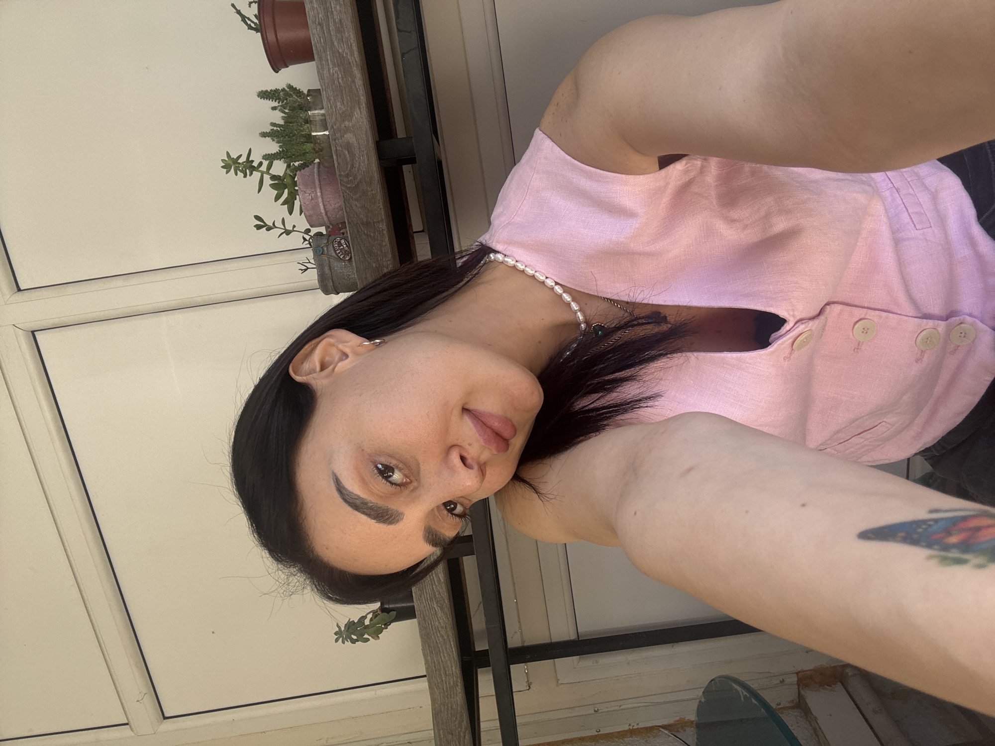 A woman takes a selfie outdoors, likely on a balcony. She is wearing a pink top, and a pearl necklace is visible. She has dark hair and fair skin. Her left arm is extended towards the camera, showing a tattoo. Behind her, there is a table with potted plants, including succulents. The backdrop includes white-framed windows. No indication of location in Tbilisi, Georgia can be determined.