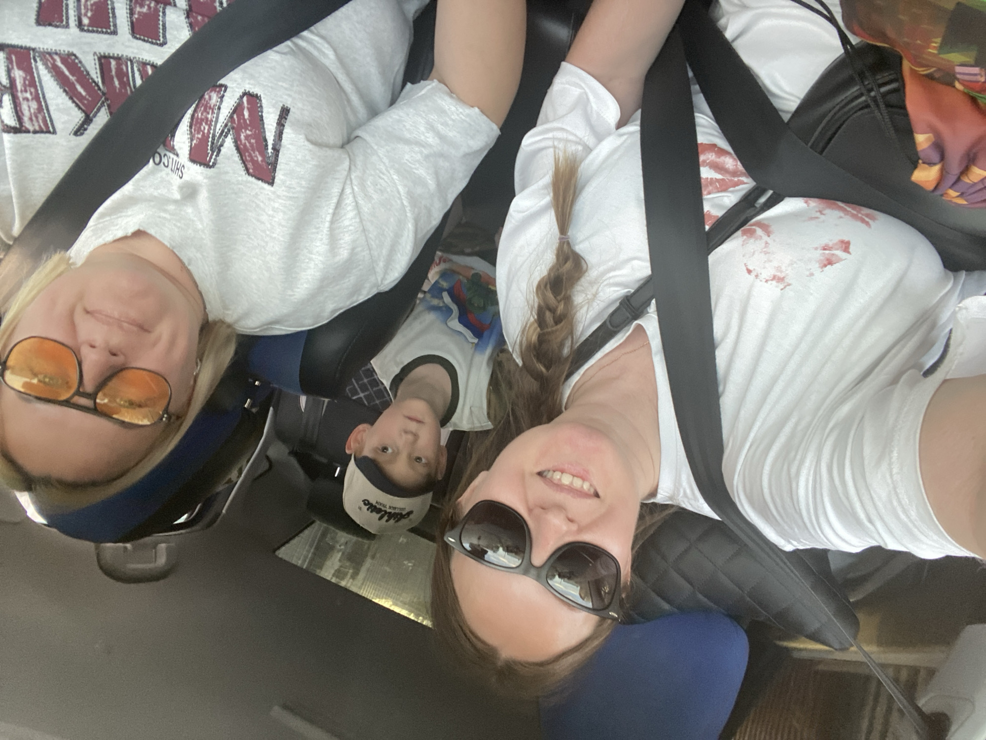 From the perspective inside a vehicle, a selfie shows three individuals. An adult female is positioned on each side of a younger male. The woman on the left has short blonde hair, wears orange-tinted glasses, and a gray shirt with a large, dark-colored inscription. The woman on the right has long brown hair styled in a braid with sunglasses, and a white t-shirt marked with what appears to be red lipstick marks. The boy in the middle has on a cap and a white t-shirt with an illustration. All are wearing seatbelts. The location is unidentifiable in the city of Chaikovsky, Russia.