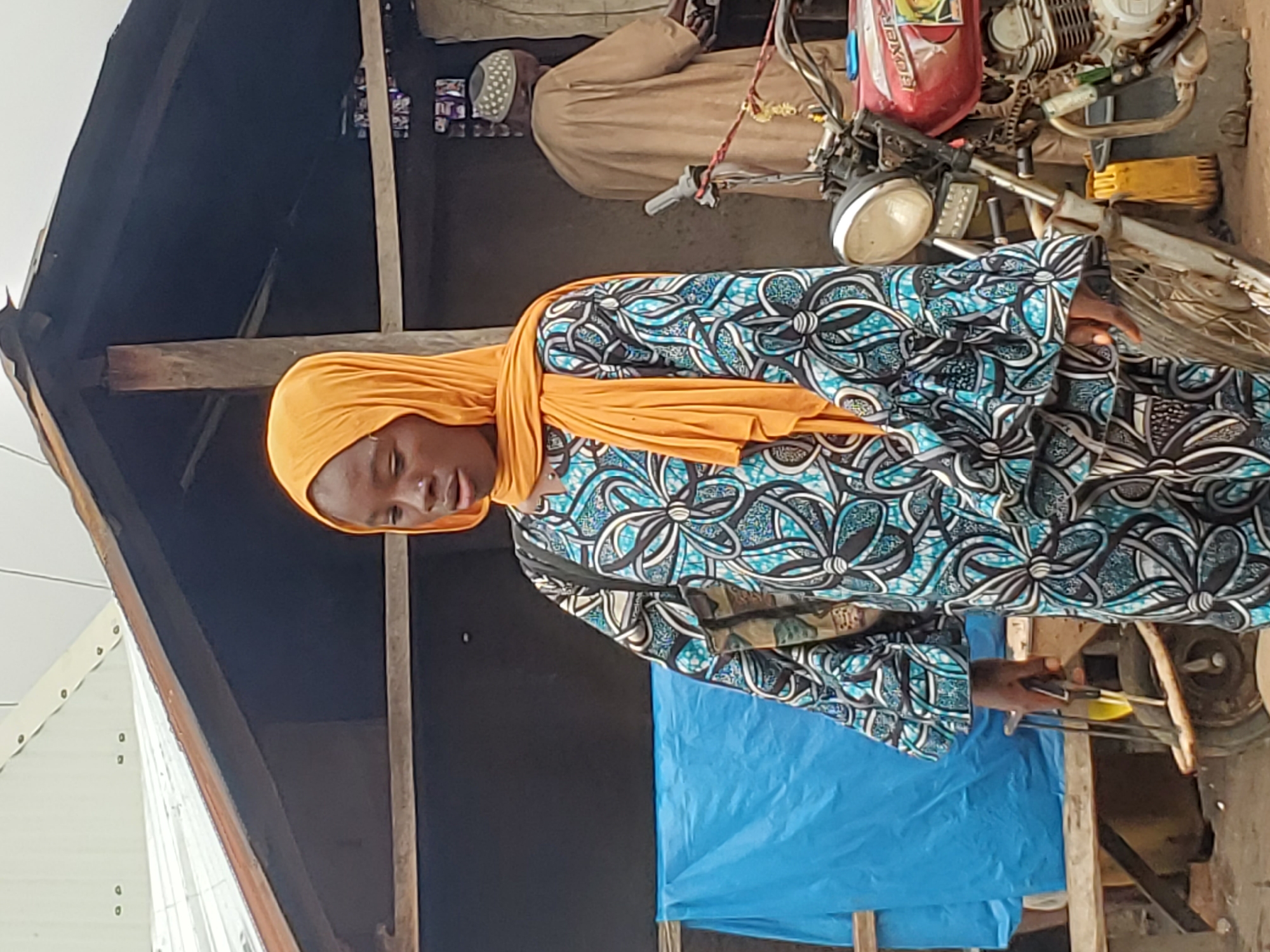 In Mararaba, Nigeria, a woman wearing a blue patterned dress and orange head covering stands outdoors, seemingly engaged in cooking with skewers or tongs. Nearby, a motorcycle is visible alongside another individual. The environment appears to be an informal outdoor setting, possibly a roadside food stall or market area. A blue tarp is also present, suggesting a makeshift structure.
