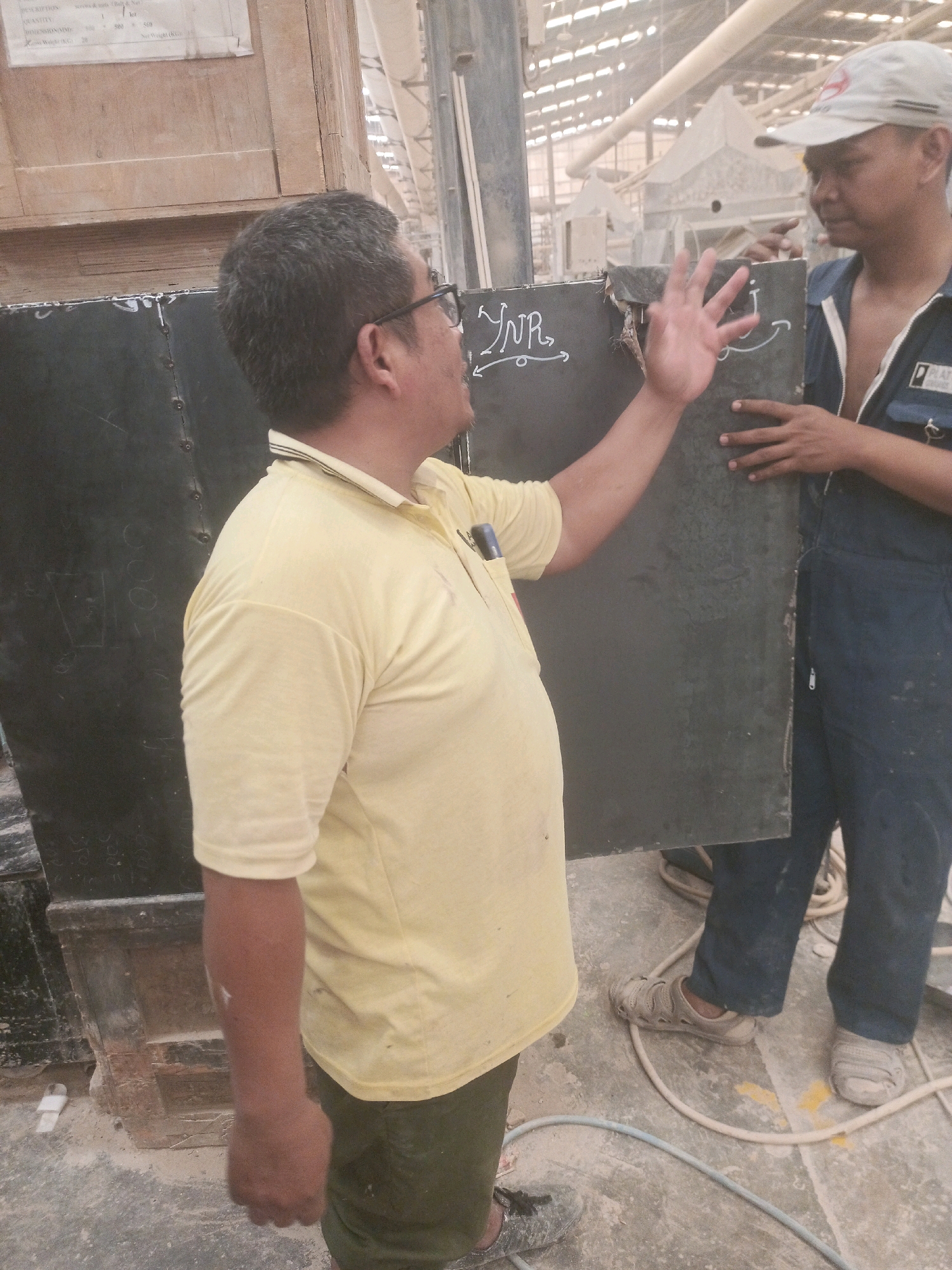 Two men are in an industrial setting, possibly a workshop or warehouse, in Lebaniwaras, Indonesia. The man on the left, wearing glasses and a yellow polo shirt, is gesturing with his left hand. A dark, rectangular object, possibly a panel or door, is positioned between the two men. The man on the right, wearing a blue uniform and a grey cap, is holding the edge of this object and looking at the other man. There are markings and writing on the dark object. The environment appears dusty, with exposed beams and industrial equipment visible in the background. Several electrical cords or hoses are present on the floor.