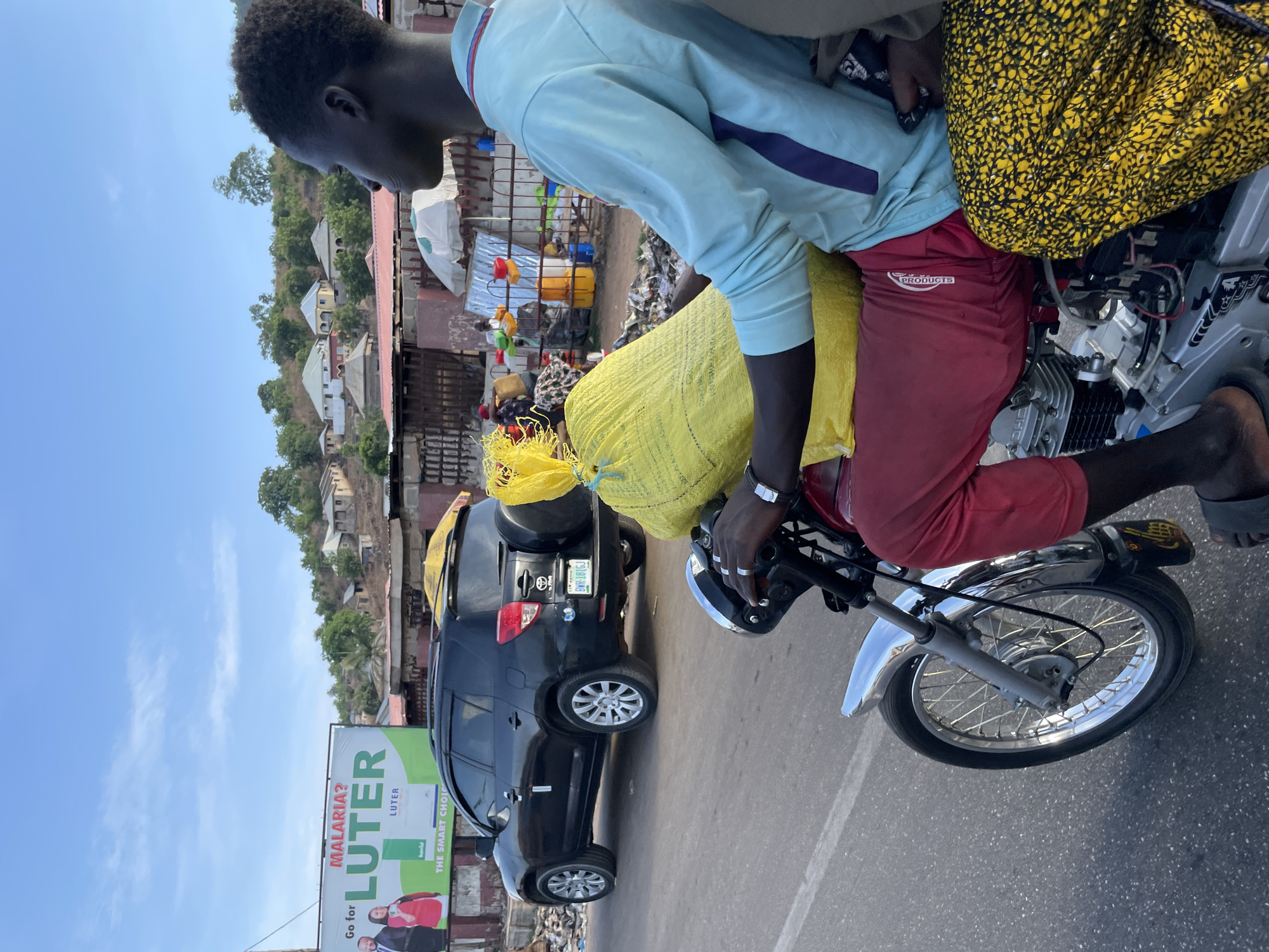 The image depicts a street scene in Lokoja, Nigeria. A man is riding a motorcycle, carrying yellow bags. He is wearing a light blue shirt and red pants. Another person is visible behind him on the motorcycle, wearing a yellow patterned top.
A black SUV is parked on the side of the road. In the background, there are buildings and structures on a hill, with vegetation visible. A billboard with the name "LUTER" is prominently displayed on the left side of the road. The sky is clear and blue.