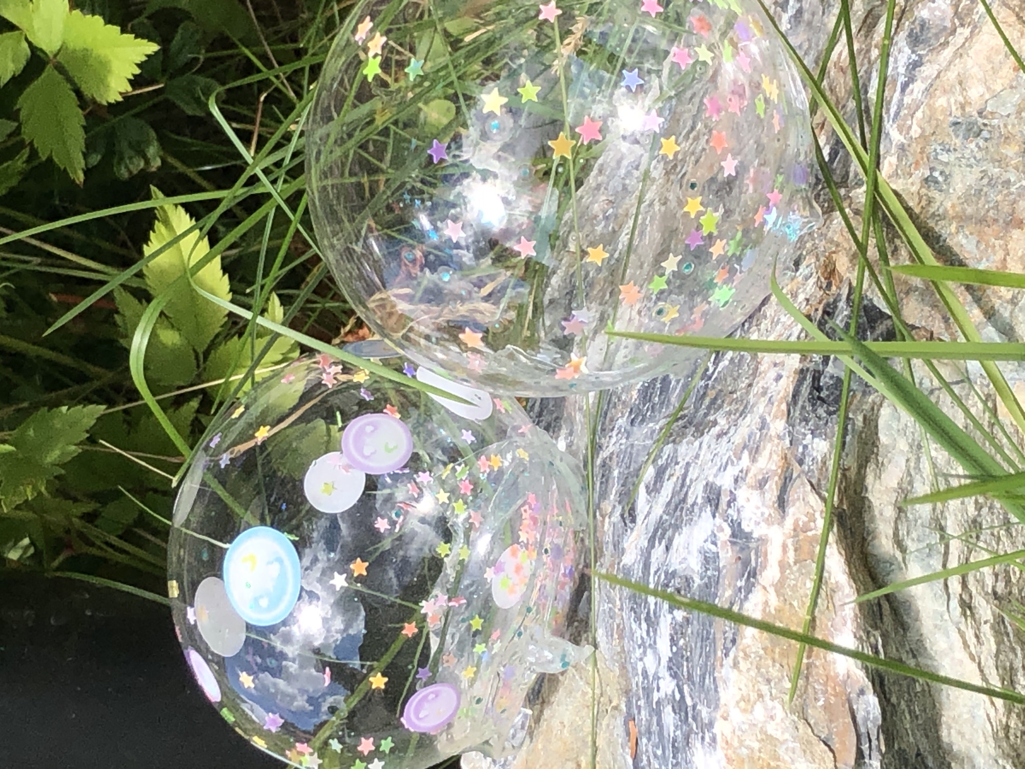Two clear, round plastic ornaments filled with colorful star-shaped confetti and small circular decals are resting on a rock formation surrounded by green grass and foliage. The ornament on the left is closer to the viewer and partially obscured by grass. It contains circular decals depicting simple smiley faces and crescent moons in pastel colors. The ornament on the right is positioned further back and to the right, with its details less clear due to distance and reflection. The background consists of lush green leaves and plants. The scene appears to be outdoors in a natural setting.