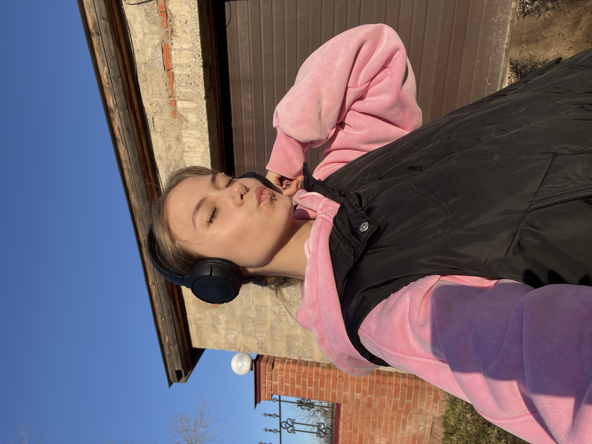 A young woman is captured in a bright, sunny outdoor setting in Фоки, Russia. She is shown from the chest up, with her eyes closed and lips pursed in a playful 'kissing' gesture. She has light-colored hair and multiple facial piercings, including one above her upper lip and two on her lower lip. Over her ears, she wears black JBL brand headphones.
She is dressed in a pink hooded sweatshirt layered under a black quilted vest. The strong sunlight casts clear shadows, indicating it is daytime. The sky above is a clear, vibrant blue. In the background, various architectural elements are visible: an old, weathered brick and concrete structure, possibly part of a house or garage, featuring a dark brown metal door or siding. Further back, another brick wall with a decorative black metal fence or gate is seen, along with a white spherical outdoor lamp. Some bare tree branches are also discernible against the sky. The overall mood conveyed by her expression and the bright light is cheerful and serene.