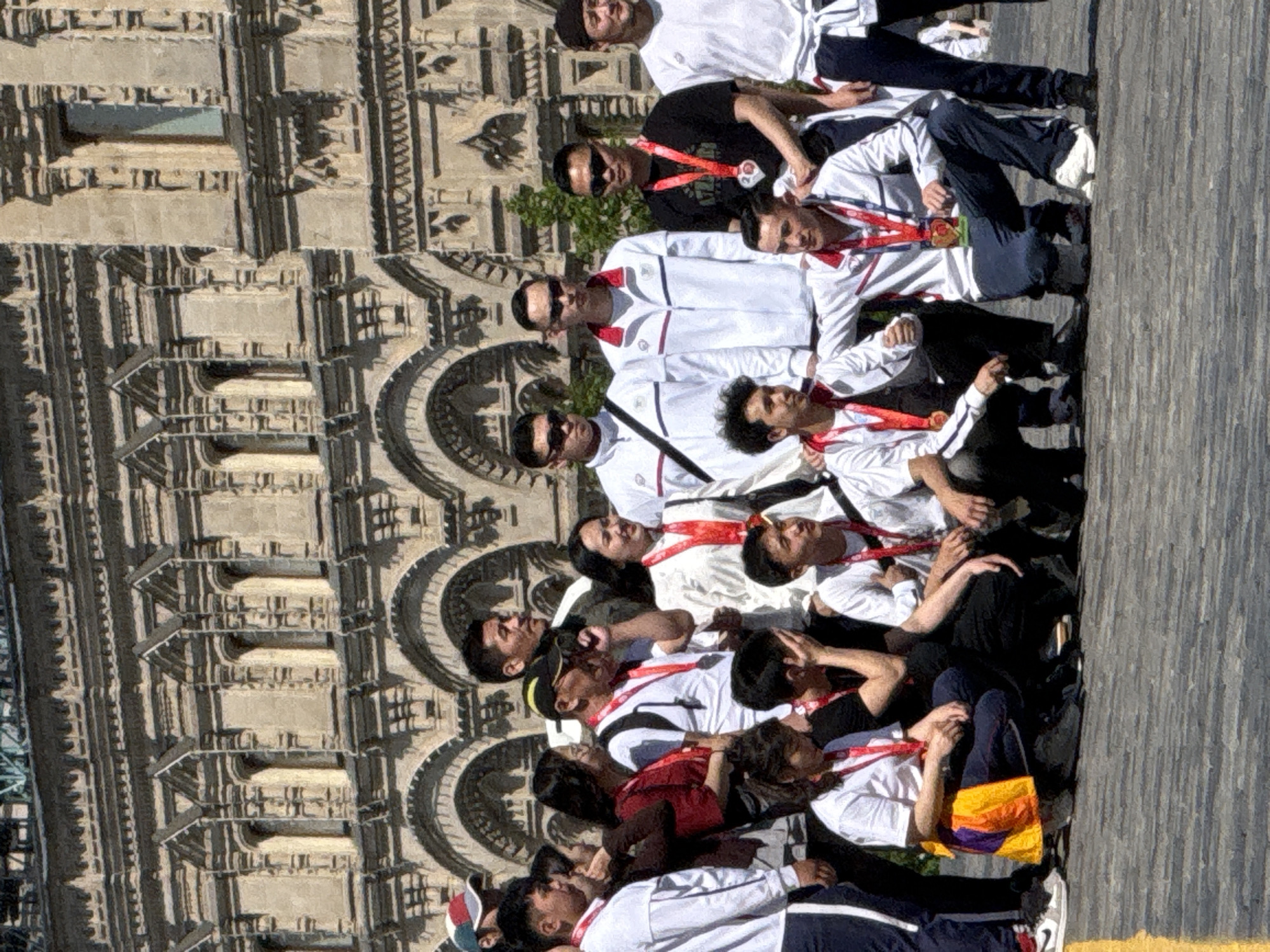 A group of approximately 15 people, mostly men, are lying on a wooden surface in front of a large, ornate building. The building features multiple levels of arched windows and intricate stonework, indicating a historical or religious structure. The group appears to be posing for a photograph or taking a rest. Many of them are wearing white shirts and tracksuits with red accents. Some individuals have medals around their necks, suggesting a recent achievement or event. Some are wearing sunglasses.
The scene is likely in Moscow, Russia, and the building in the background may be St. Basil's Cathedral or a similar landmark in the city center.