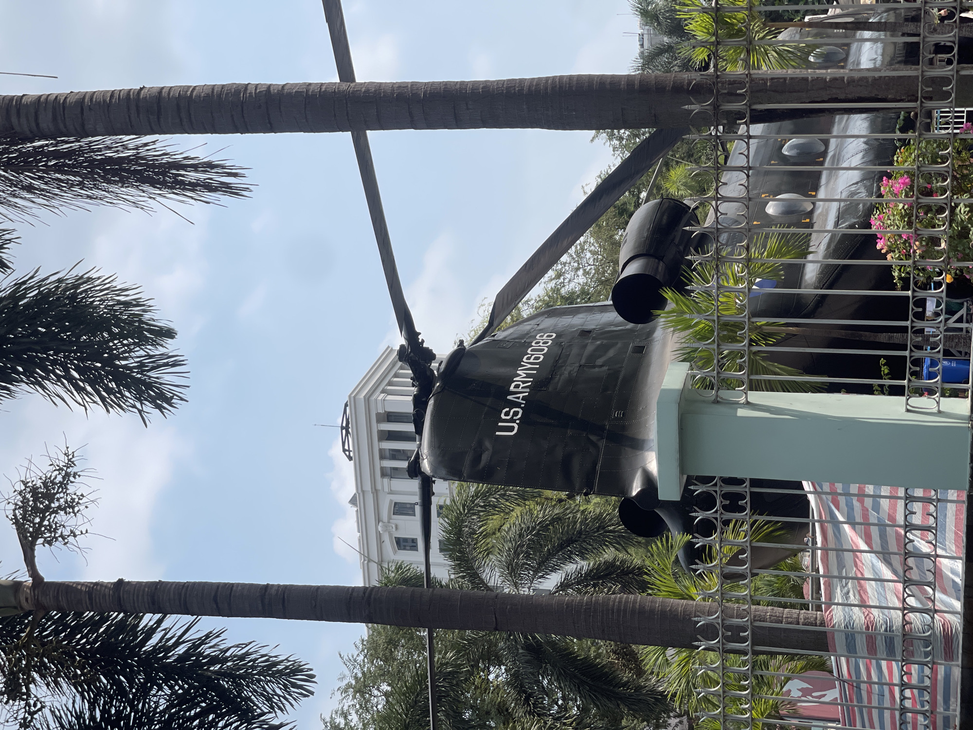 A partial view of a U.S. Army helicopter with the designation "U.S. ARMY 6086" is visible through a metal fence in Thủ Đức, Vietnam. The helicopter is mounted on a pedestal. Palm trees are interspersed in the foreground and background. In the background, a white building is partially visible. The sky is pale blue with light clouds.