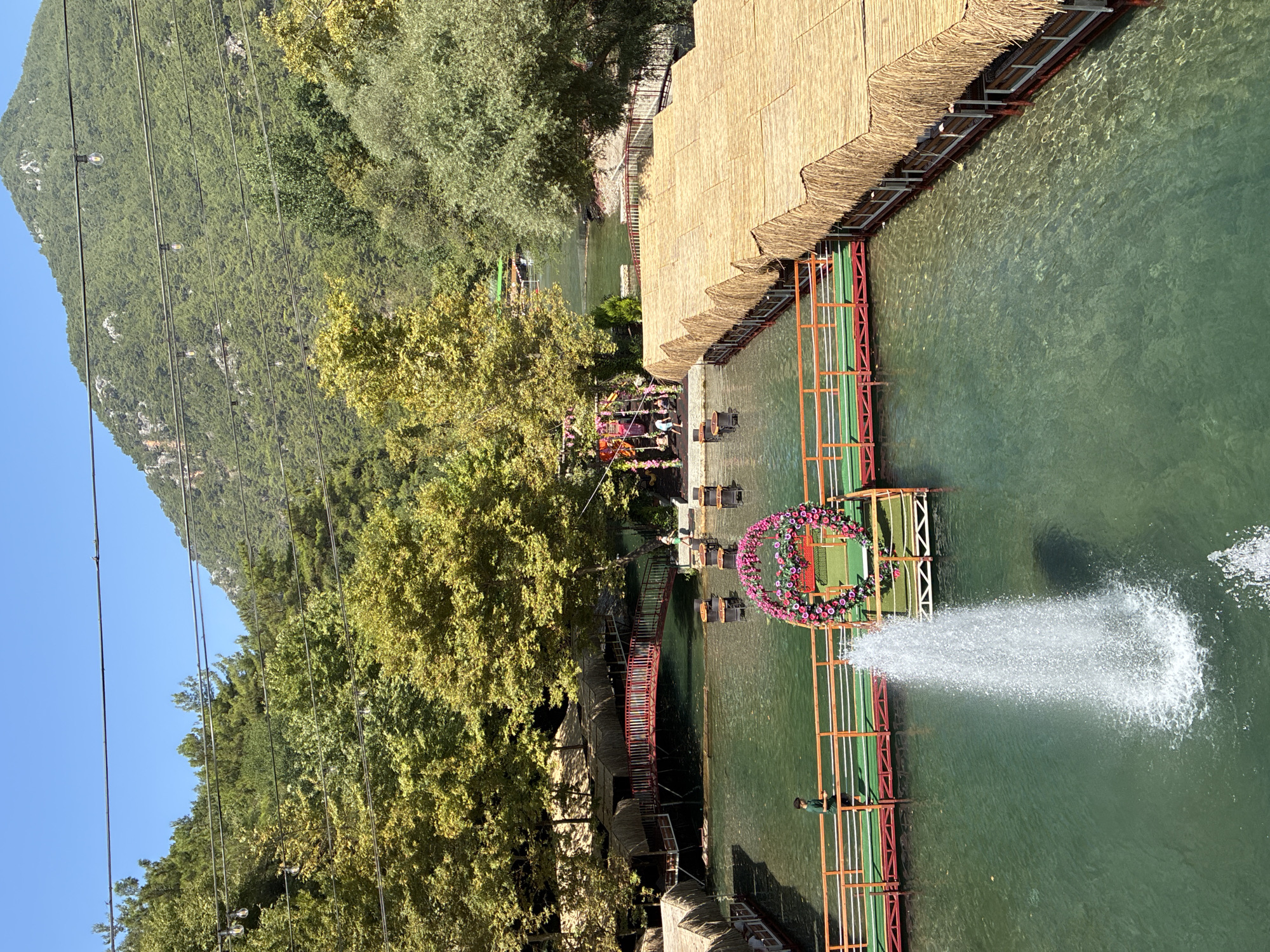 The image depicts a serene water park or resort area situated in Alanya, Turkey, nestled at the base of a lush, tree-covered mountain under a clear blue sky. The foreground features a tranquil, emerald-green body of water with a central fountain creating a dynamic spray. Several elevated wooden walkways with red and green railings crisscross the water, leading to thatched-roof dining platforms that extend from the shore. One walkway supports a vibrant floral archway, likely a decorative element. In the mid-ground, a charming red arched bridge spans the water, connecting different sections of the resort. A few individuals are visible, including a person walking along a walkway and another near the red bridge, suggesting the area is operational and frequented by visitors. The background is dominated by dense green foliage, with trees of varying shades of green and yellow, indicative of a temperate climate. The mountain slopes are heavily forested, creating a natural backdrop. The overall atmosphere is one of natural beauty and recreational tranquility.