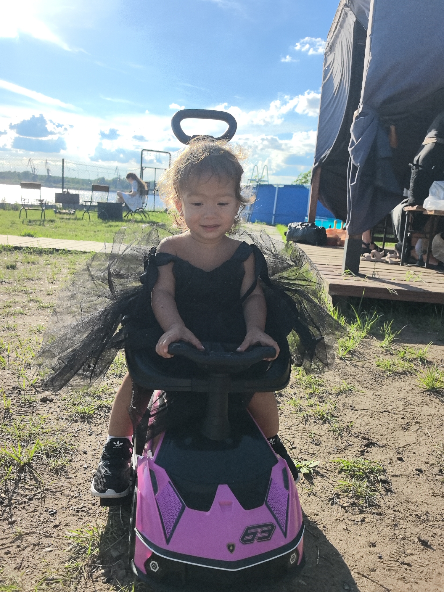 A young girl, appearing to be a toddler, sits on a pink toy car. She is dressed in a black tutu and dress, with a black headband that resembles angel wings. She is smiling and looking down at the steering wheel of the toy car. The car has a black steering wheel and a pink body with black accents and a silver "63" emblem. The girl is wearing black athletic shoes with white soles and laces.
The setting is outdoors on a sunny day. The ground is dirt and grass. In the background, there is a body of water with industrial cranes along the distant shore. There are also some chairs and what appears to be a picnic area. To the right of the girl, there is a dark tent-like structure. The sky is blue with scattered white clouds. The sunlight suggests it is daytime, likely afternoon given the shadows. The location context indicates Chaikovsky, Russia. There is no specific event or activity clearly depicted beyond the girl sitting on the toy car.