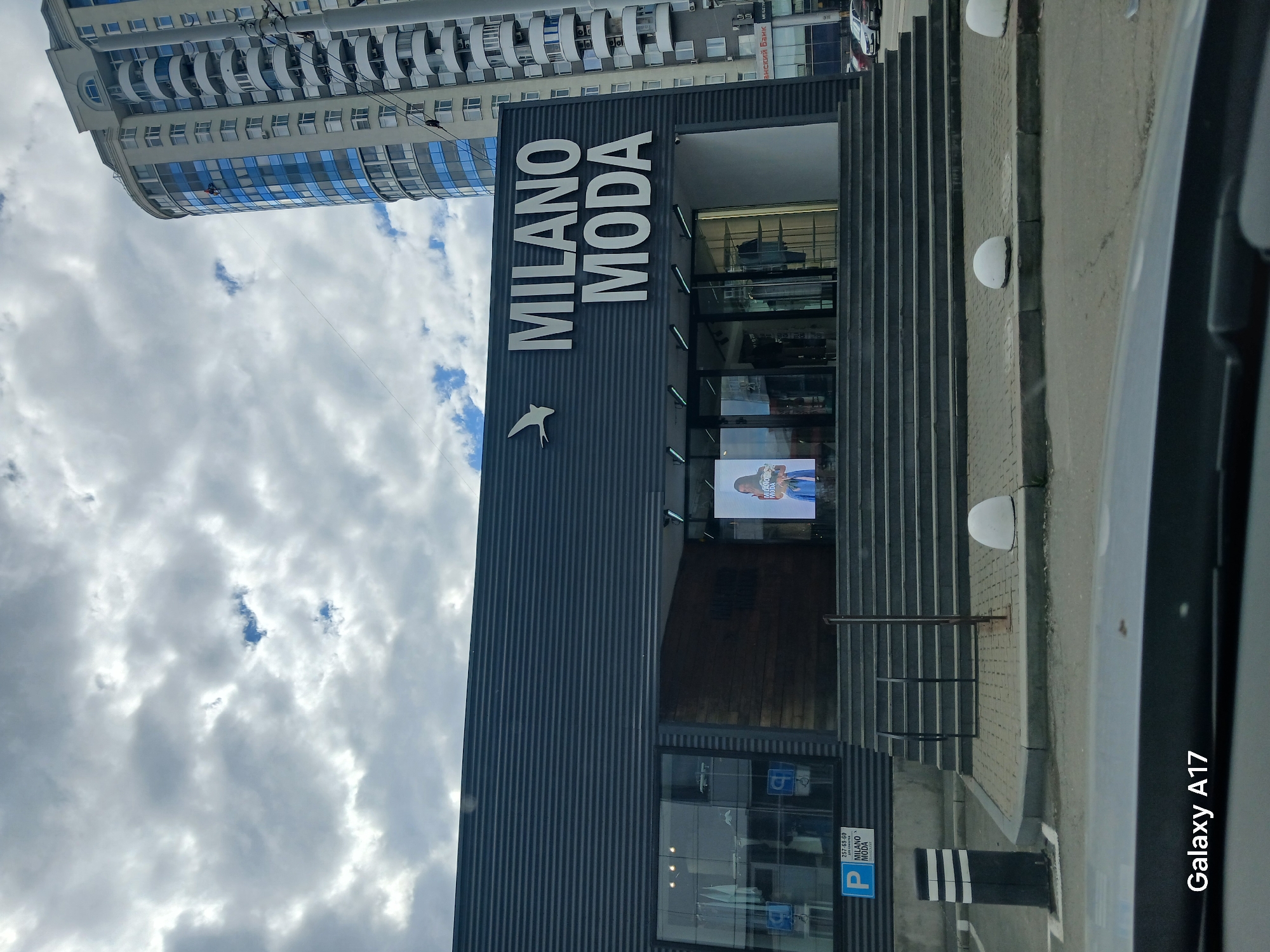 The image shows the exterior of the Milano Moda store in Yekaterinburg, Russia. The building has a modern design, with a dark gray facade featuring vertical stripes. The store's name, "MILANO MODA," is displayed prominently in large white letters. A bird-like symbol is also visible on the facade. A set of stairs leads to the store's entrance. A tall, modern building is visible in the background. The sky is cloudy. A car is partially visible on the right side of the image. A parking sign with the store's name and phone number is present.