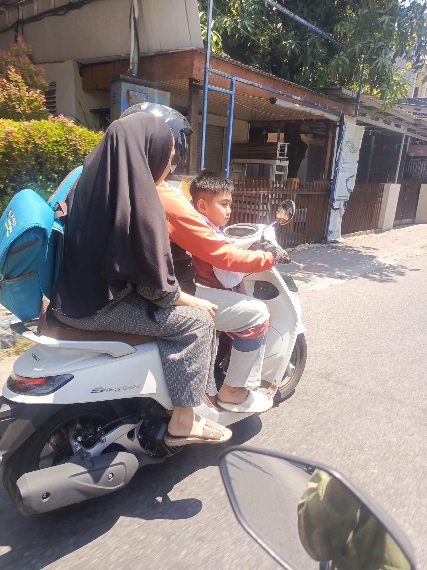This is a medium shot, taken from a slightly elevated angle, of two people riding a white scooter on a paved road in Bebekan, Indonesia. The person in the back is a woman wearing a black hijab and a black headscarf, a black top, and grey striped pants. She has a blue backpack on her back. The person in front is a young boy wearing a red and orange long-sleeved shirt and grey pants with red and blue stripes. He is wearing gloves and is holding onto the handlebars of the scooter. The scooter has the word "HONDA" and "Style" visible on it. In the background, there is a building with a wooden fence and trees. The sun is shining, casting shadows on the road. The image is slightly blurry, suggesting movement.