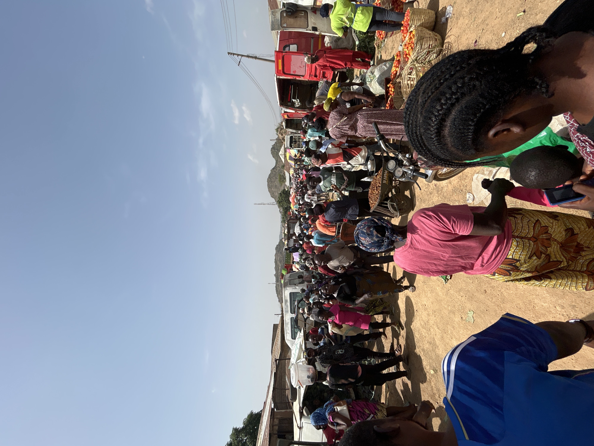 The image shows a crowded marketplace in Bukuru, Nigeria. The scene includes multiple trucks, some red and some white, which seem to be used for transporting goods. People are densely packed, possibly engaged in buying or selling produce, as baskets of tomatoes are visible near one of the trucks. Individuals are wearing varied clothing, with a mix of colors and patterns. A mountain is visible in the background. The ground is dusty and dry. A motorcycle is parked near the forefront of the crowd, and its carrier is loaded with what appear to be dried fruits or nuts.