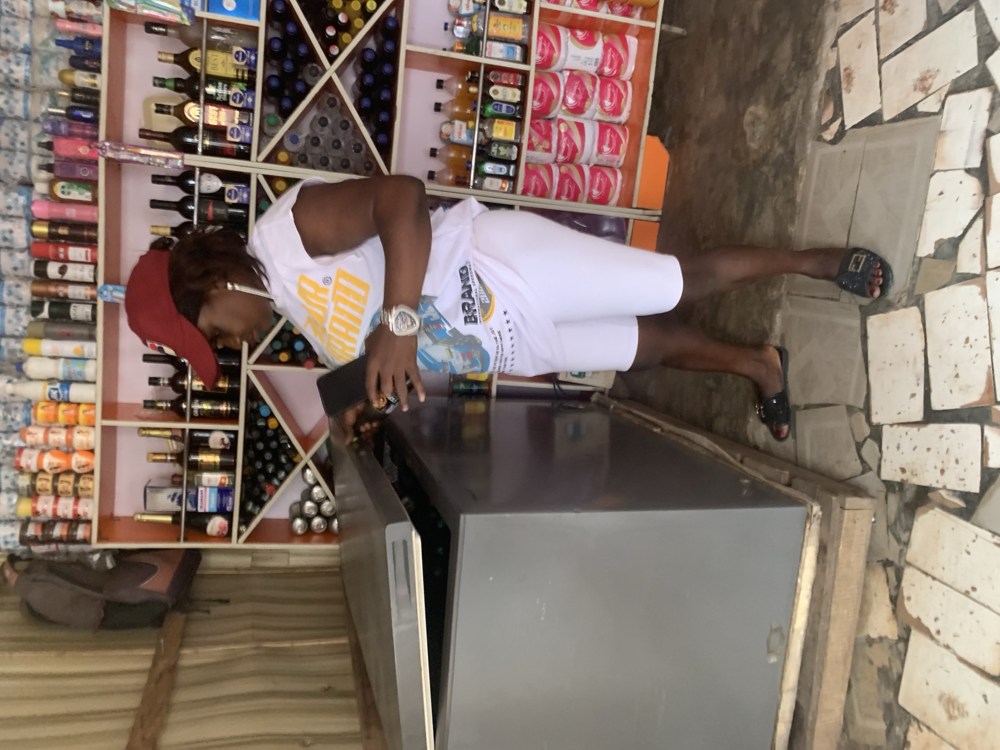 In Shagamu, Nigeria, a woman stands next to an open freezer, holding a dark rectangular object, likely a mobile phone. She wears a red baseball cap, a white t-shirt with yellow lettering, and white shorts, along with black sandals. The freezer is gray and appears to be in a store or market setting. Behind her, shelves are stocked with various items, including bottles of alcohol, canned goods, and toiletries. The floor is tiled with a pattern of light and dark stones. The walls are covered with corrugated metal sheeting.