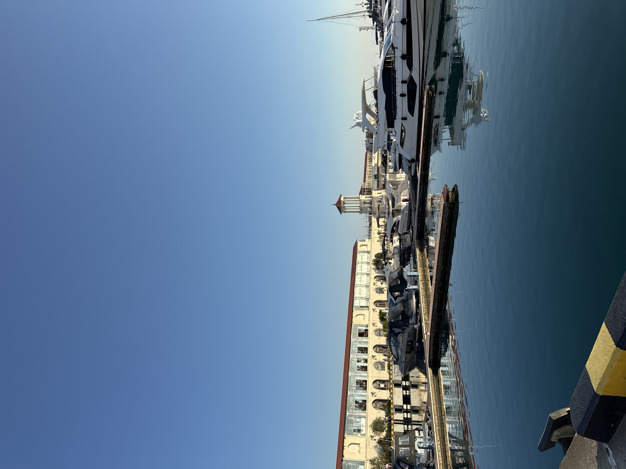 The image depicts a marina in Sochi, Russia, on a clear, sunny day. The sky is a vibrant blue, taking up the left half of the frame. The right half is dominated by the calm, reflective surface of the water, which mirrors the scene along the quay.
Moored along the quay are several luxury yachts of varying sizes, their sleek white hulls contrasting with the dark water. In the background, a multi-story building with a red-tiled roof and a light-colored facade lines the waterfront. This building features numerous windows and arched doorways, giving it a distinct architectural style. A prominent tower with a reddish-brown roof is visible, extending above the building.
The activity taking place is the mooring of these yachts in a bustling marina. The overall atmosphere appears serene and prosperous, suggesting a leisurely or vacation setting. The time of day is likely midday or early afternoon, given the bright sunlight and the clear sky. The reflection of the building and yachts in the water adds a sense of depth and tranquility to the scene.
Visible text is limited to a black and yellow striped bollard in the foreground, which serves as a marker for the dock. The boats themselves may have some branding or hull numbers, but these are not clearly legible at this resolution. The image captures the essence of a high-end maritime location, highlighting the interplay between architecture, water, and luxury vessels under a bright, open sky.