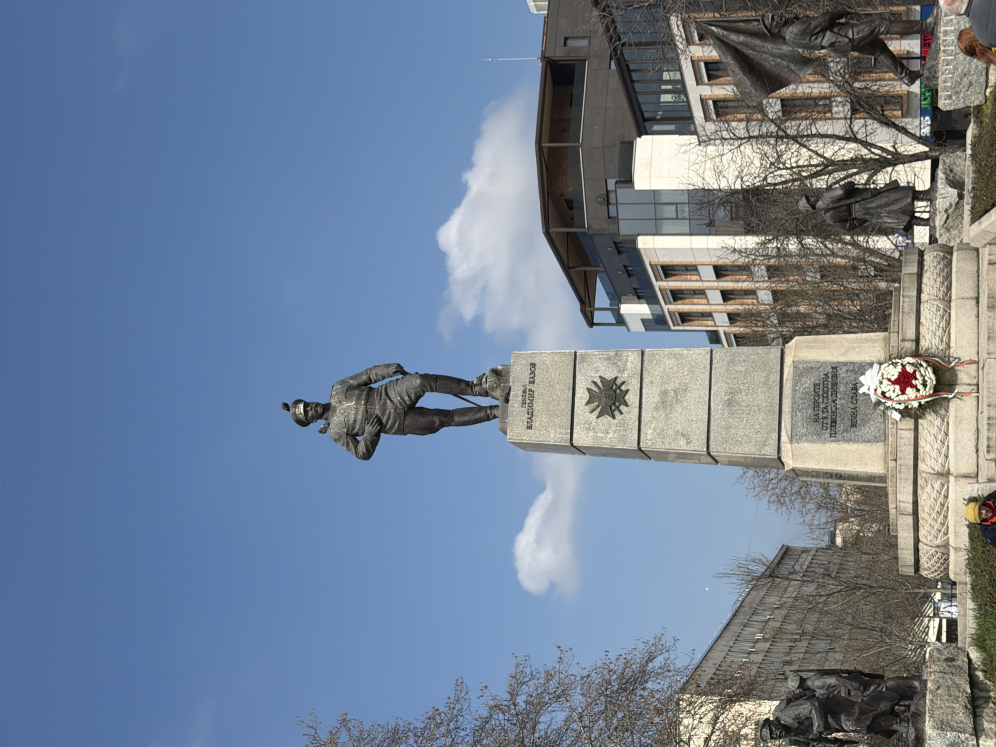 The image shows the monument of General Vladimir Vazov in Pleven, Bulgaria. The bronze statue of General Vazov stands on a high, rectangular granite pedestal. The pedestal features inscriptions and a metallic decoration. In the background, there's a blue sky with some clouds, along with the facades of modern buildings. In the bottom right, there are parts of a sculpture and a building with some people. The scene appears to be captured during daylight hours.