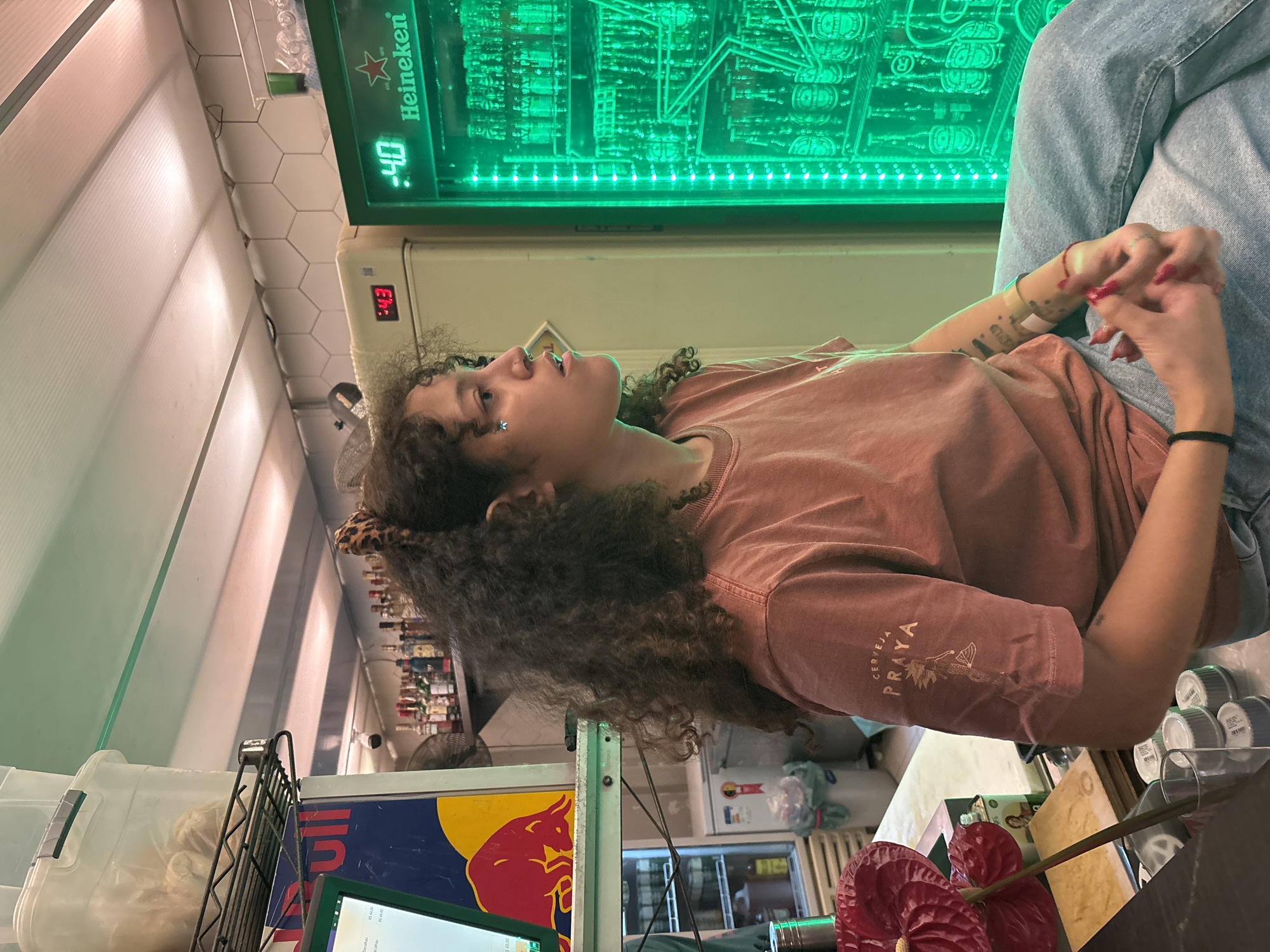 This image, captured in São Paulo, Brazil, depicts an indoor scene within what appears to be a bar or commercial establishment. The primary subject is a young person with dark, curly hair, adorned with facial piercings (nose and lip) and a star-shaped earring, with a leopard print hair accessory partially visible. They are wearing a reddish-brown t-shirt, which displays "CERVEJA PRAIA" (Beach Beer) and a beach-themed graphic on the sleeve, paired with light blue jeans. Their hands are clasped, revealing red nail polish, and there's a bandage or tape on one arm over faint tattoos. The person gazes upwards and slightly to the right, presenting a contemplative or observant expression.
In the background, a prominent green-lit Heineken display or cooler is visible, showing "Heineken" text and a digital reading of ":40". Below it, another cream-colored refrigerator or cooler displays ":43". White hexagonal tiles line a wall to the left. The counter area in the foreground features a Red Bull logo, a partially visible computer screen showing what resembles a spreadsheet, a clear plastic container, and two vibrant red anthurium flowers. Further back, shelves stocked with various liquor bottles and a fan confirm the bar-like setting. The artificial lighting suggests an indoor evening environment.