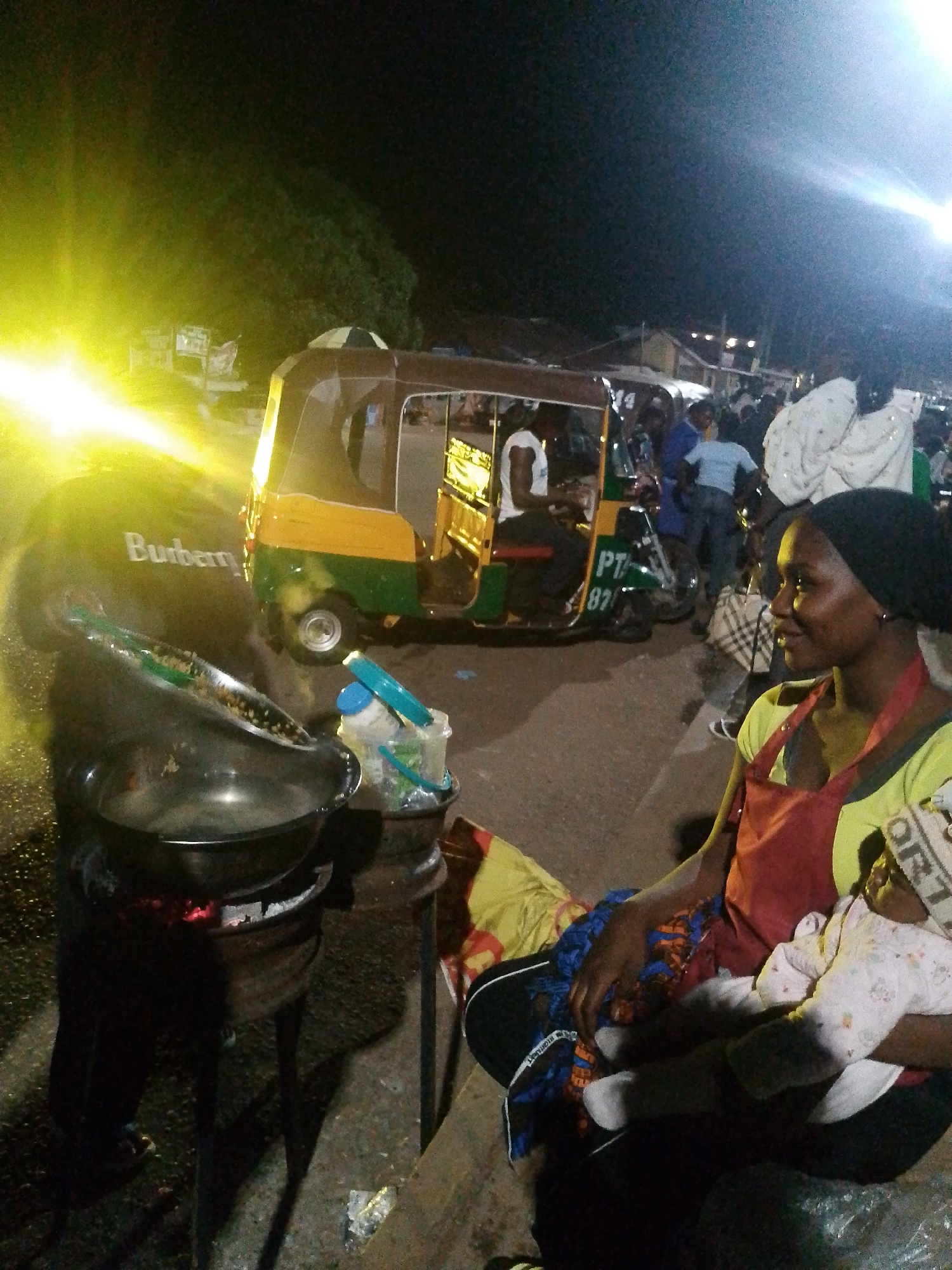 The image depicts a nighttime scene in an outdoor urban setting, possibly a street market or roadside area. In the foreground, a woman wearing a black headscarf, a yellow shirt, and a red apron is holding a baby. The baby is swaddled in a light-colored outfit with a pattern. Behind the woman and to her left, a cooking setup is visible, featuring a large metal pot placed over a makeshift stove with glowing embers. There's also a bucket with items inside.
In the mid-ground, a colorful auto-rickshaw, painted yellow and green with the text "PT 87" on its side, is parked. Several people are standing around the auto-rickshaw and in the background, suggesting a busy environment. The lighting in the scene is a mix of artificial light sources, including a bright, intense yellow light emanating from the left side of the frame, possibly from a vehicle's headlights or a strong lamp. The background is dark, with some distant structures and trees visible. The ground appears to be paved. The overall atmosphere suggests a lively commercial or social gathering at night.