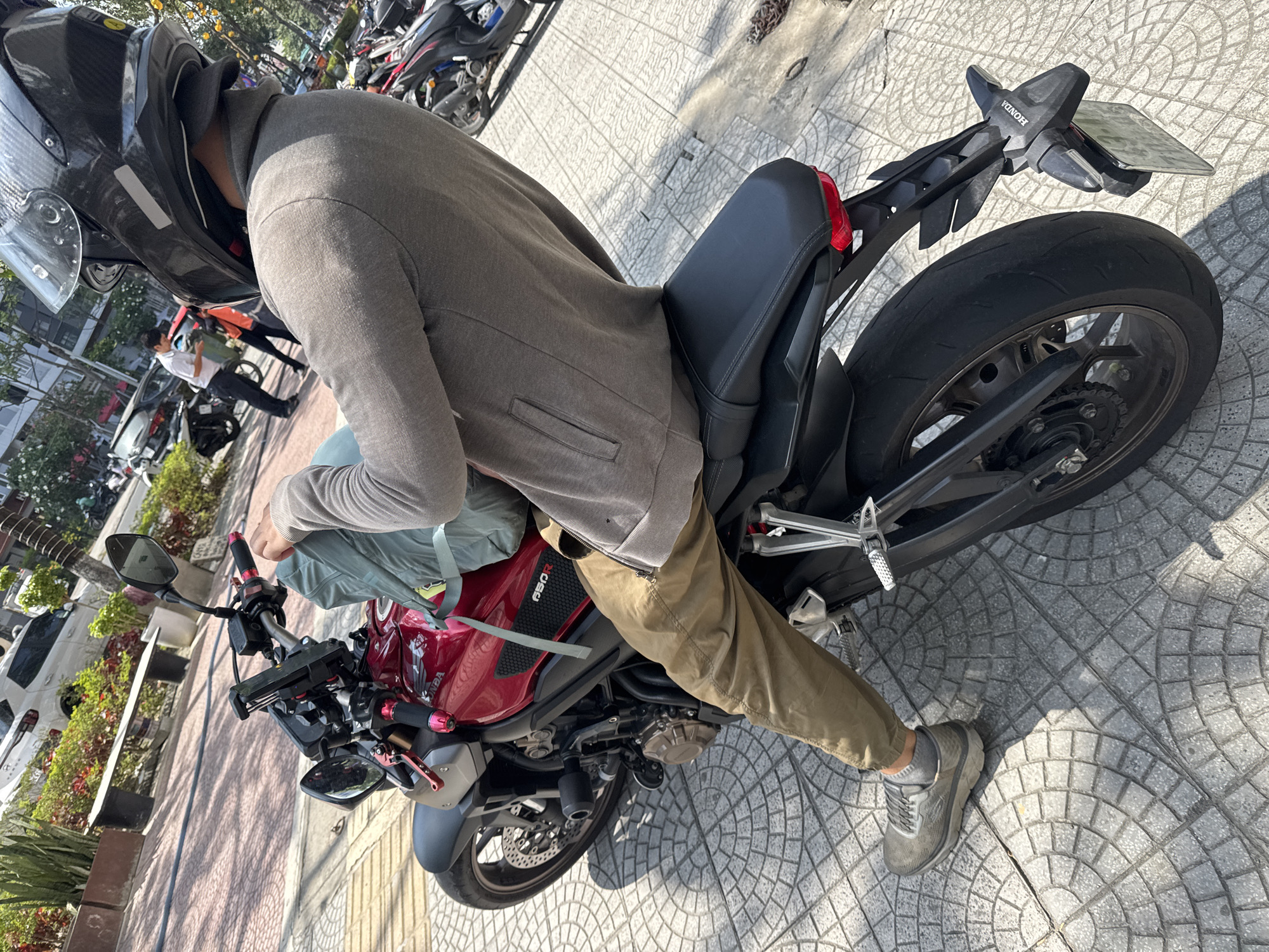 A person wearing a helmet, gray jacket, and khaki pants is sitting on a red Honda CB650R motorcycle on a sidewalk in Đà Nẵng, Vietnam. The sidewalk is paved with gray tiles. The motorcycle is partially obscured by the person. Other motorcycles are parked in the background. Trees and buildings are visible in the background, suggesting an urban environment. The lighting indicates it is daytime.
