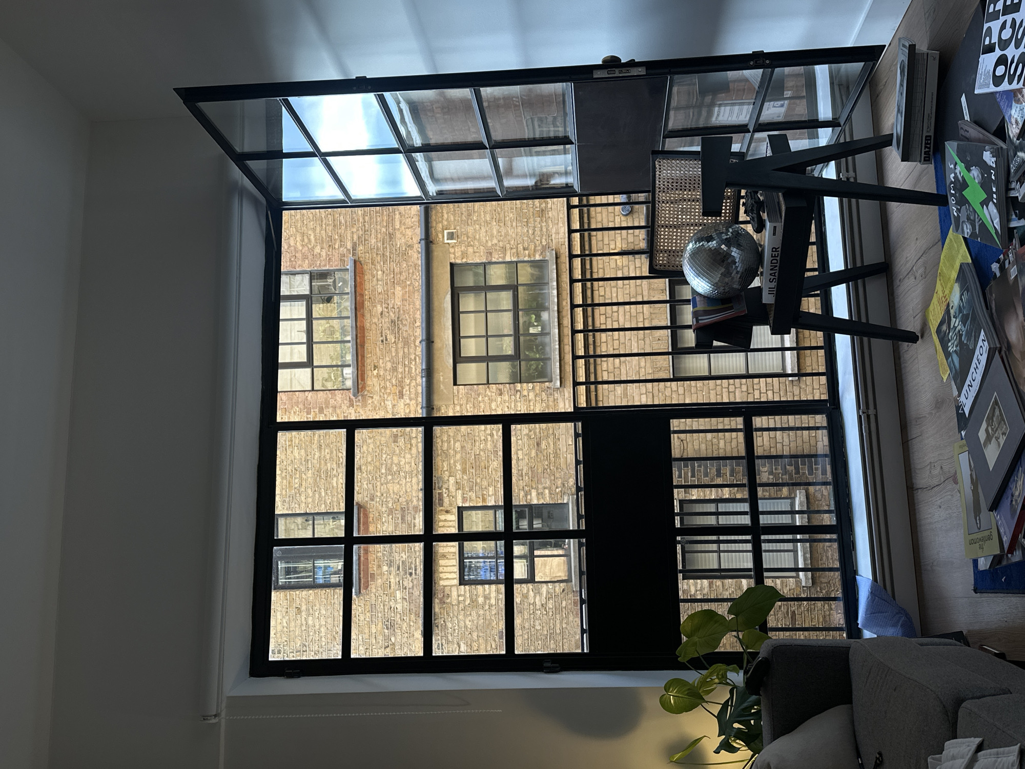 The image shows an interior view, looking out through a large industrial-style window with black frames. The view is of an adjacent brick building with multiple windows. Some of the windows have darker frames and different sizes. The window frame in the foreground features a small glass roof at the top. To the right of the window, there is a modern table with a wicker chair, books, a disco ball, and various magazines, including one titled "DAZED" and another featuring "JIL SANDER." Parts of a grey couch and a houseplant are also visible in the foreground, towards the bottom right corner. The lighting suggests it's either an overcast day or the scene is primarily illuminated by interior lighting. The location is likely to be London, United Kingdom, based on the architectural style and design features often associated with converted industrial buildings in London.