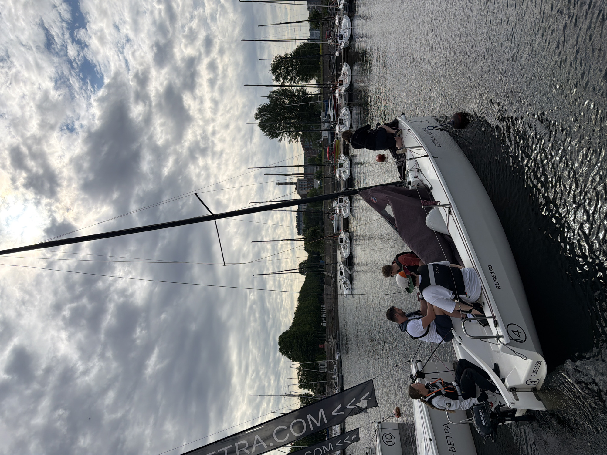 This is a medium-angle, eye-level shot of several sailboats docked on a body of water, likely the Neva River in Saint Petersburg, Russia, under a cloudy sky. In the foreground, a white sailboat is partially visible, with several people on board. A woman wearing a white jacket and life vest is seated on the left side, looking forward. Next to her, a man in a white t-shirt and life vest is seated. Another man in a white t-shirt and life vest is also seated. To the right, a person in a black jacket is kneeling on the boat. The boat has the registration number "RUS6169" and the number "4" visible. Another white sailboat with the number "10" is to the left. In the background, a line of sailboats are docked, with trees and buildings visible beyond them. The water is dark and choppy. Two large black flags with white text are positioned on the left side of the frame, with text that includes "TRA.COM<<" and "COM<<".