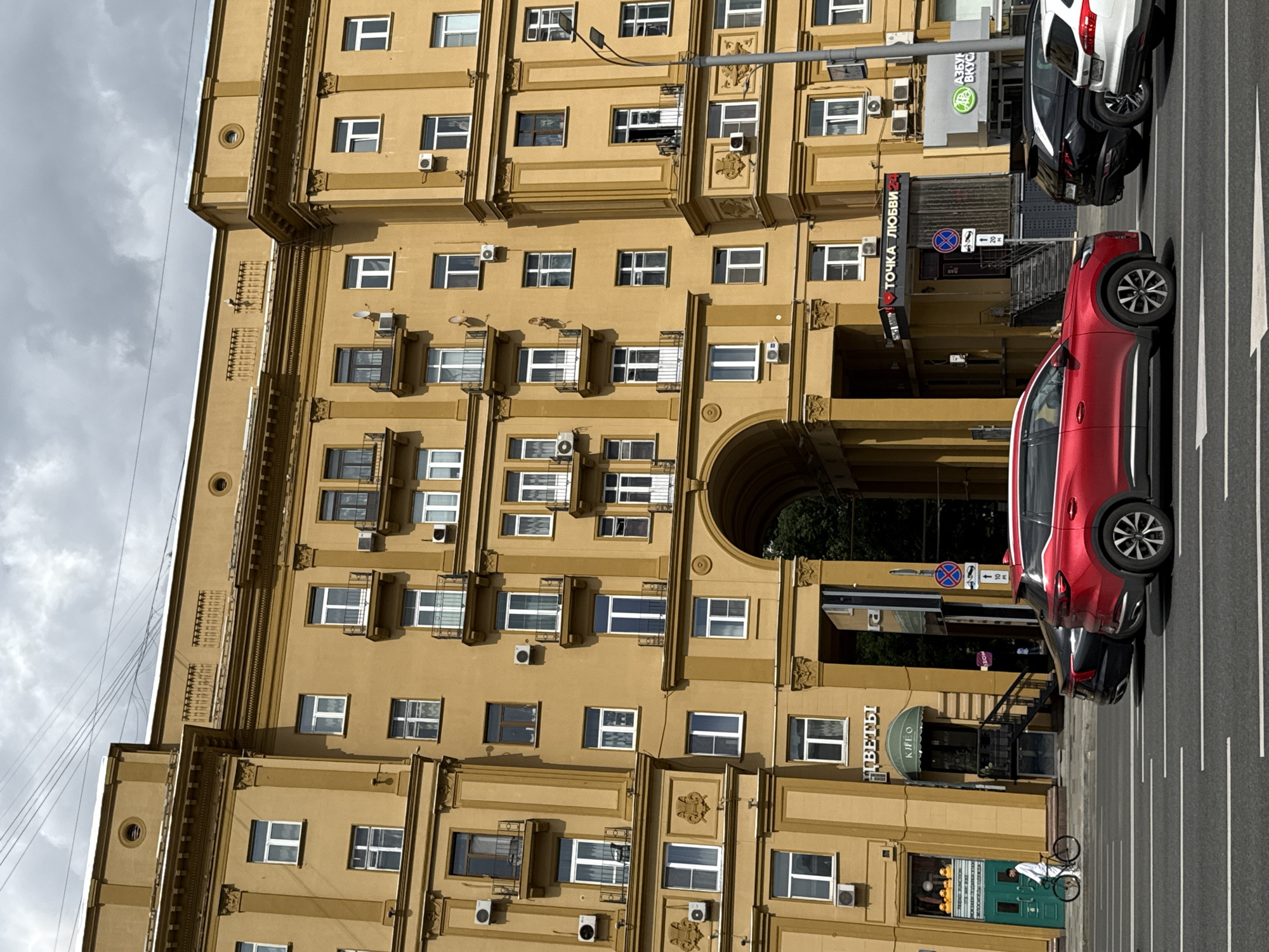 The image shows a building in Moscow, Russia, with a street in front of it. The building is tall and yellow with many windows and decorative elements. Several cars are parked on the street, including a red SUV and a black car. There is a bicycle parked in front of a green door. The sky is cloudy. Text visible on the building includes "ТОЧКА ЛЮБВИ 24" and "АЗБУКА ВКУСА". The overall impression is of an urban scene on a cloudy day.