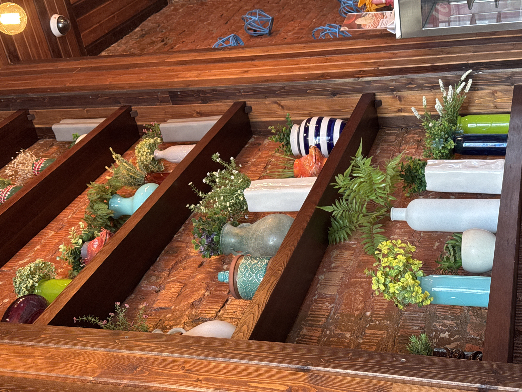 This image captures a decorative wall display within an indoor setting, likely a restaurant or cafe in Birobidzhan, Russia. The scene features a series of wooden shelves and beams arranged diagonally, set against a textured background of reddish-brown brickwork and additional wooden paneling.
The shelves are adorned with a variety of decorative objects, primarily vases and bottles of different shapes, sizes, and colors, including light blue, green, white, striped blue and white, teal, purple, and red-orange. Many of these containers hold artificial plant arrangements or small flower sprigs in white, yellow, and pink hues, adding touches of greenery and floral accents to the display.
In the upper part of the frame, several abstract, blue geometric cage-like sculptures are visible on a higher wooden surface. To the far left, a warm-toned light bulb in a fixture and a white dome-shaped security camera are mounted on a wooden beam, indicating the commercial nature of the space. Part of a metallic display case or appliance is also visible in the upper right corner.
No human activity is taking place; the image is a static shot of an artfully arranged decorative installation. The lighting appears to be artificial and warm, contributing to a cozy or inviting atmosphere. The textures of the brick, wood, and various decorative items are prominent. No visible text is present in the image.