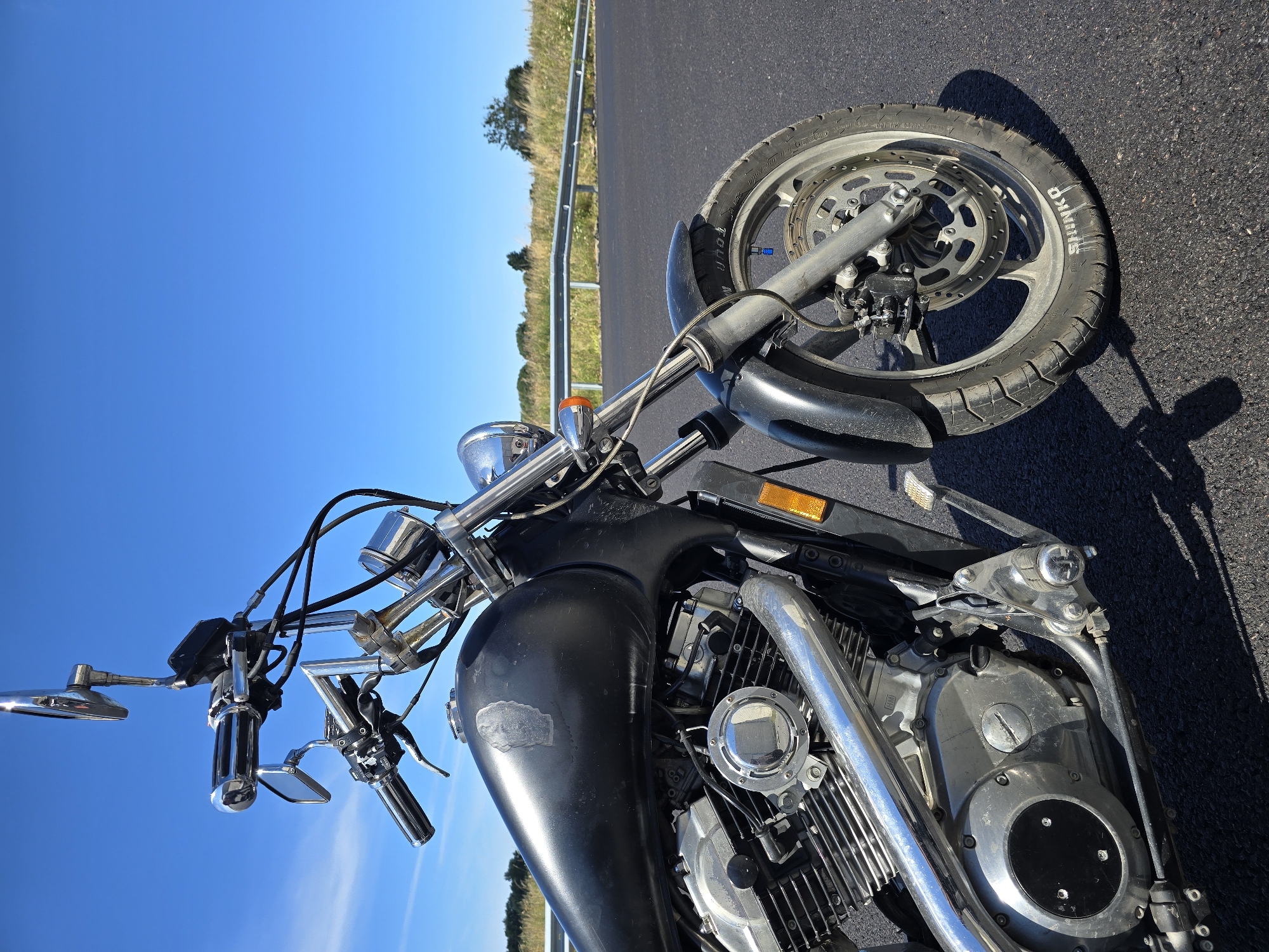 A black motorcycle is parked on an asphalt road. The motorcycle is angled with the front wheel on the right side of the frame. The handlebars, chrome accents, engine, and part of the fuel tank are visible. The motorcycle is in the foreground, with a grassy embankment and a guardrail in the mid-ground. The background consists of a clear blue sky with a few wispy clouds and distant trees. There are no people visible in the image. The scene appears to be a roadside rest stop or pull-off. The image was taken in the city of Listovoe, Russia.