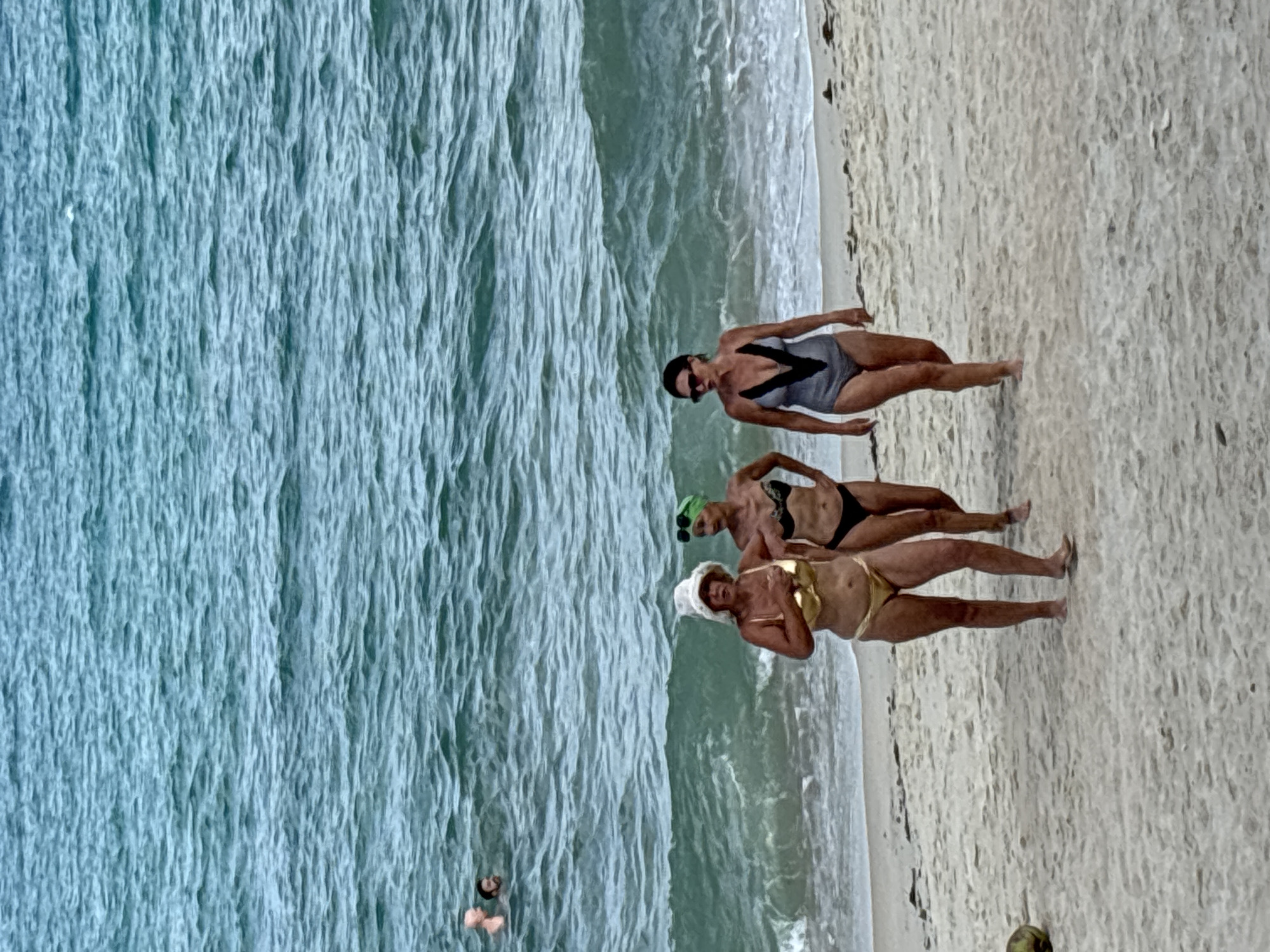 The image shows a beach scene. Three women in swimwear are standing on the sand near the water's edge. A fourth person is seen swimming in the water. The location is presumed to be Baan Hua Thanon, Thailand. The environment is a beach setting with sand and ocean waves.