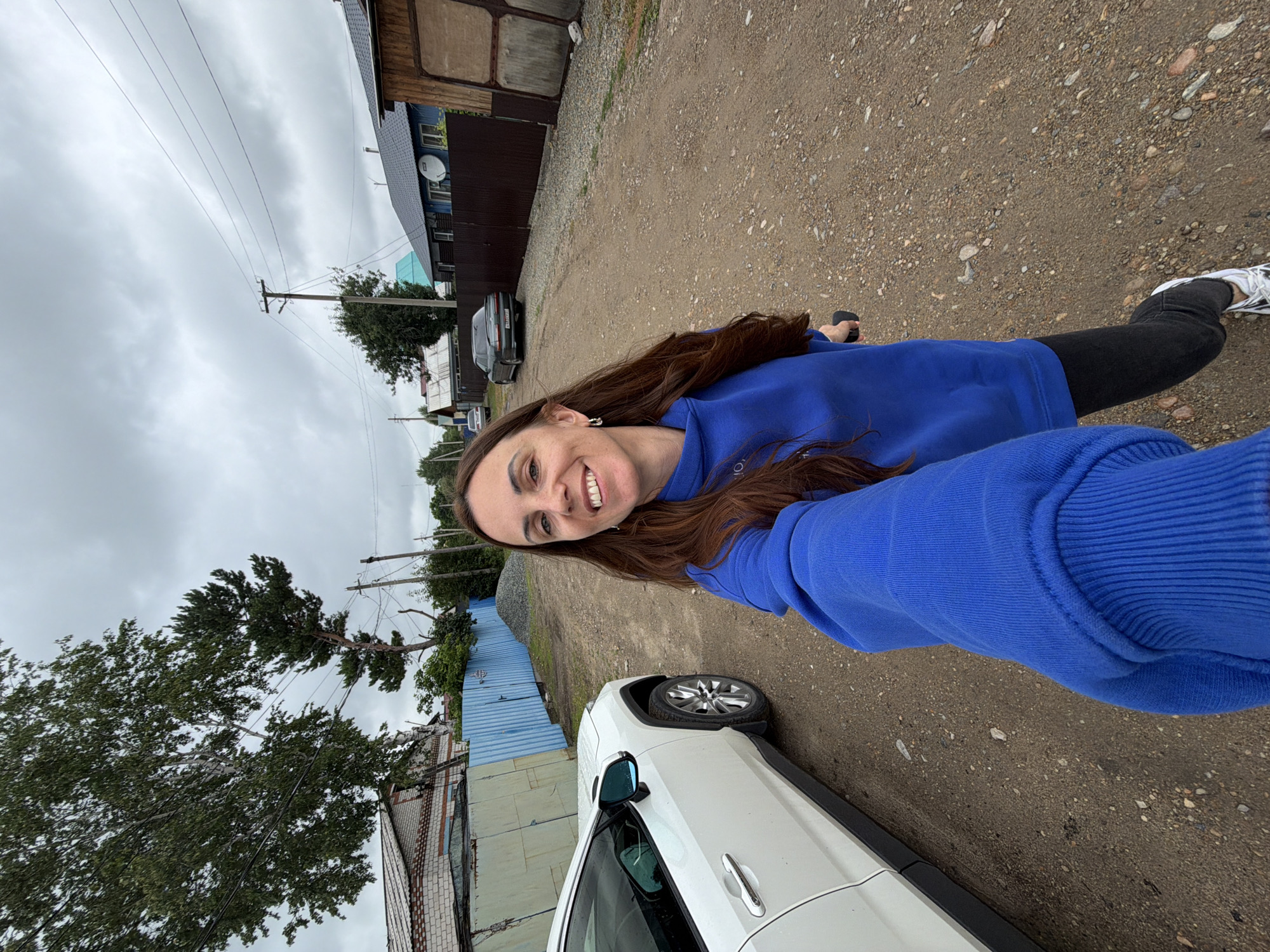 In Ptichnik, Russia, a woman with long brown hair takes a selfie outdoors on a cloudy day. She is wearing a bright blue hoodie, black pants, and white sneakers. A white car is parked to her left, and a dirt road stretches out behind her, lined with houses and fences. The scene suggests a residential area, possibly a village or rural outskirts, with elements of everyday life such as vehicles and homes.