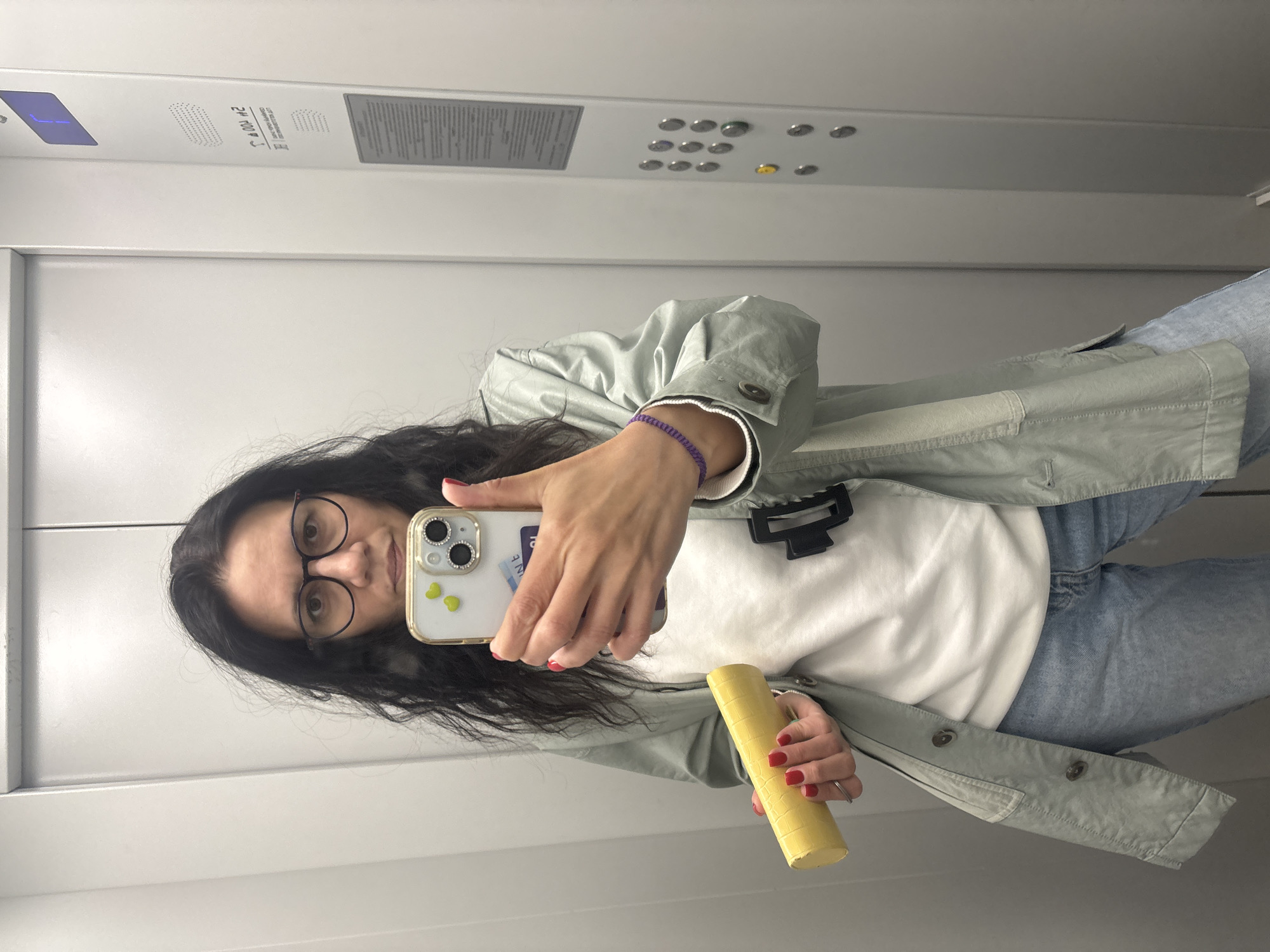 A woman with dark hair and glasses is taking a selfie in what appears to be an elevator. She is holding a yellow cylindrical object in her left hand. She is wearing a light green jacket over a white shirt and blue jeans. Her phone has a clear case with small green heart stickers. The elevator has a metal panel with buttons and display. The location is not discernible as Chaikovsky, Russia.