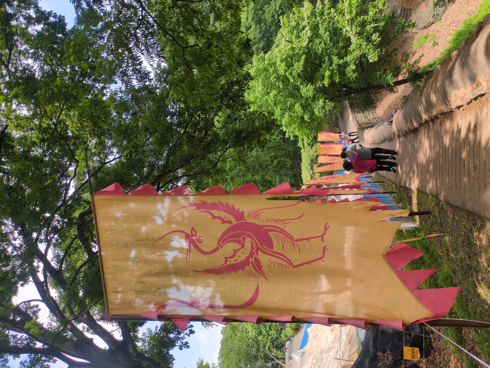 A partially obscured, long, narrow, medium shot depicts a sunny, tree-lined pathway bordered by yellow fabric flags with a red, mythical bird design, likely located in Gongju-si, South Korea.
The flags are strung overhead, creating a festive atmosphere along the path. The path, likely a walking trail, is wide and paved. Groups of people are seen walking in the distance, the closest appearing to be a couple. The surrounding environment features lush, green trees, suggesting a park or wooded area. In the foreground, a flag with a jagged, red border dominates the scene, displaying a faded red image of a phoenix-like creature. Beneath the flags, and to the side of the path, a black fence with yellow warning signs are visible.