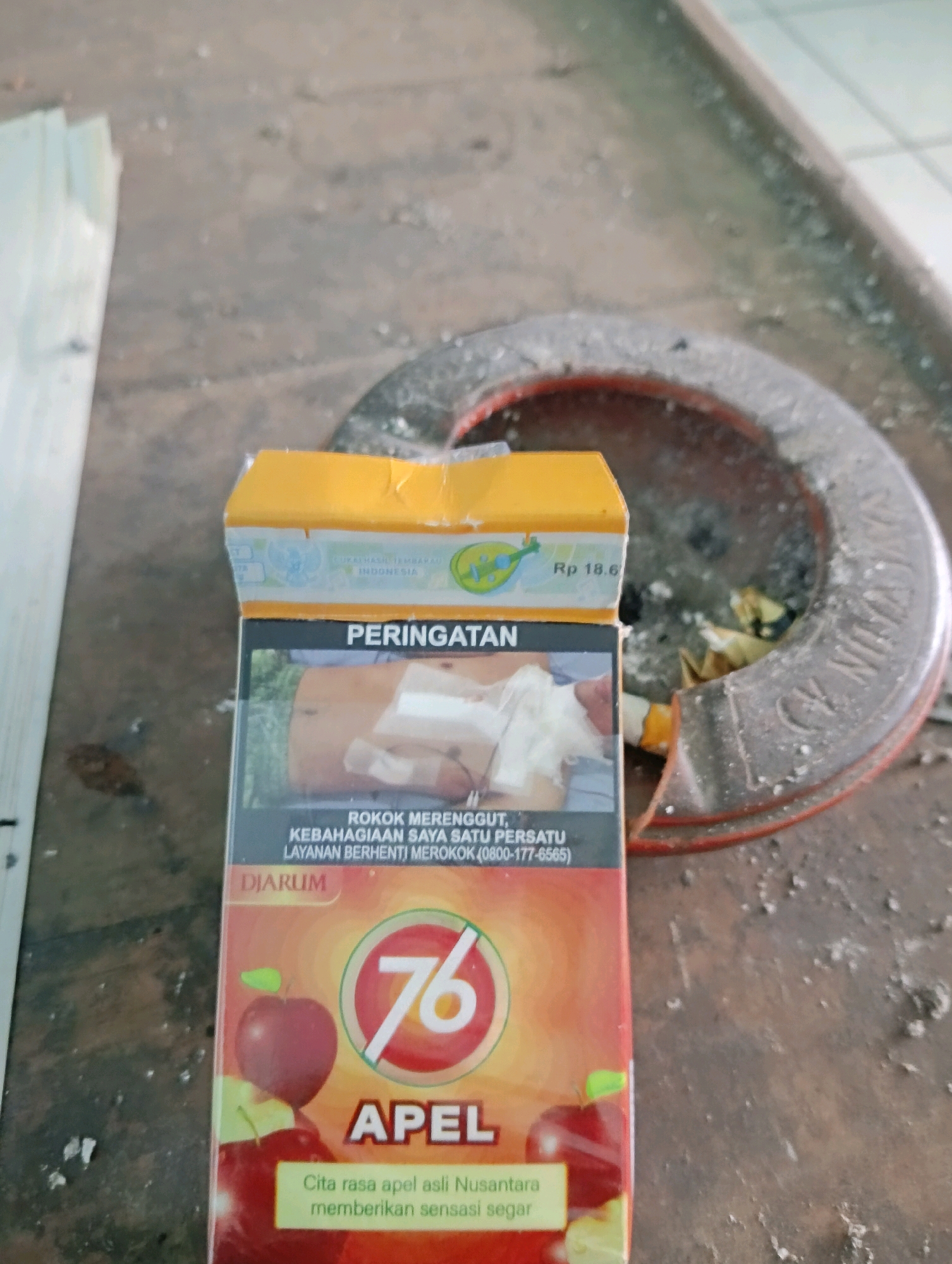 A pack of "76 APEL" cigarettes is placed on a textured, greyish-brown surface. The pack is predominantly orange and red, featuring images of apples and the number "76" in a white circle. Text in Indonesian is visible on the pack, including "PERINGATAN" (Warning) and "ROKOK MERENGGUT KEBAHAGIAN SAYA SATU PERSATU LAYANAN BERHENTI MEROKOK" (Smoking steals my happiness one by one, Quit Smoking Service). A price of "Rp 18.6" is also visible. To the right of the cigarette pack is a round, ashtray-like object filled with cigarette butts and ash. The background is out of focus but appears to be a rough, outdoor surface, possibly concrete or dirt, with scattered debris. The lighting is natural, casting soft shadows. The scene is devoid of any people or animals. The image composition suggests the pack and ashtray are the primary subjects. The location is identified as Sokaraja Kidul, Indonesia.