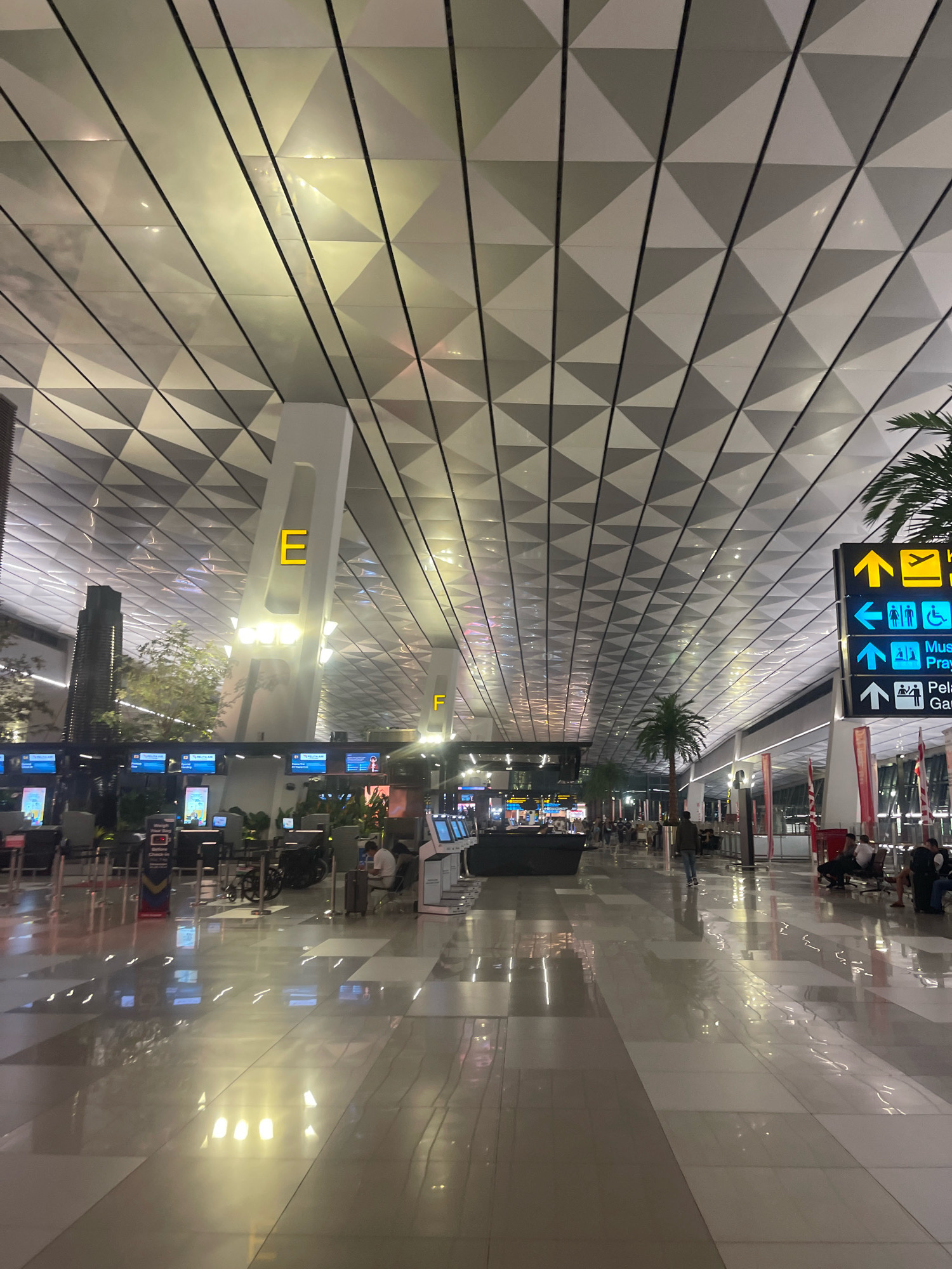 The image captures an interior view of a modern airport terminal, likely at night given the lighting. The ceiling features a distinctive geometric pattern of triangular panels, with integrated lighting that reflects off the polished floor. A large white pillar with the letter "E" in yellow is prominently displayed. In the background, check-in counters with digital screens are visible, along with a few scattered palm trees. A directional signpost with arrows in yellow and blue indicates various facilities, including "Mus" (possibly for Museum or Musholla/prayer room) and "Pelayanan" (service), with icons for restrooms and accessibility. Several individuals are present, some seated on benches, while others walk through the spacious concourse. The overall atmosphere suggests a busy, well-lit, and contemporary airport environment. The location is specified as the city of Pajang, Indonesia.