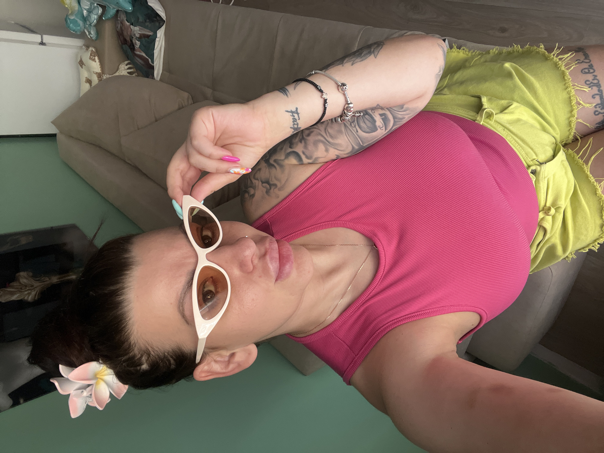 The image shows a woman with dark hair styled in a bun and adorned with a pink and white flower. She is wearing white cat-eye sunglasses with orange lenses, a bright pink ribbed tank top, and lime green shorts with frayed edges. The woman has tattoos on her arms and legs, as well as bracelets on her left wrist and multiple rings on her fingers, some of which are brightly colored. She is posing for a selfie, looking at the camera and adjusting her sunglasses. The background is an indoor setting with a muted green wall on the left and a beige sofa on the right, on which some pillows and other objects are visible. The lighting suggests it is daytime. The location is not specified in the image or its context, and therefore cannot be determined to be Plovdiv, Bulgaria.