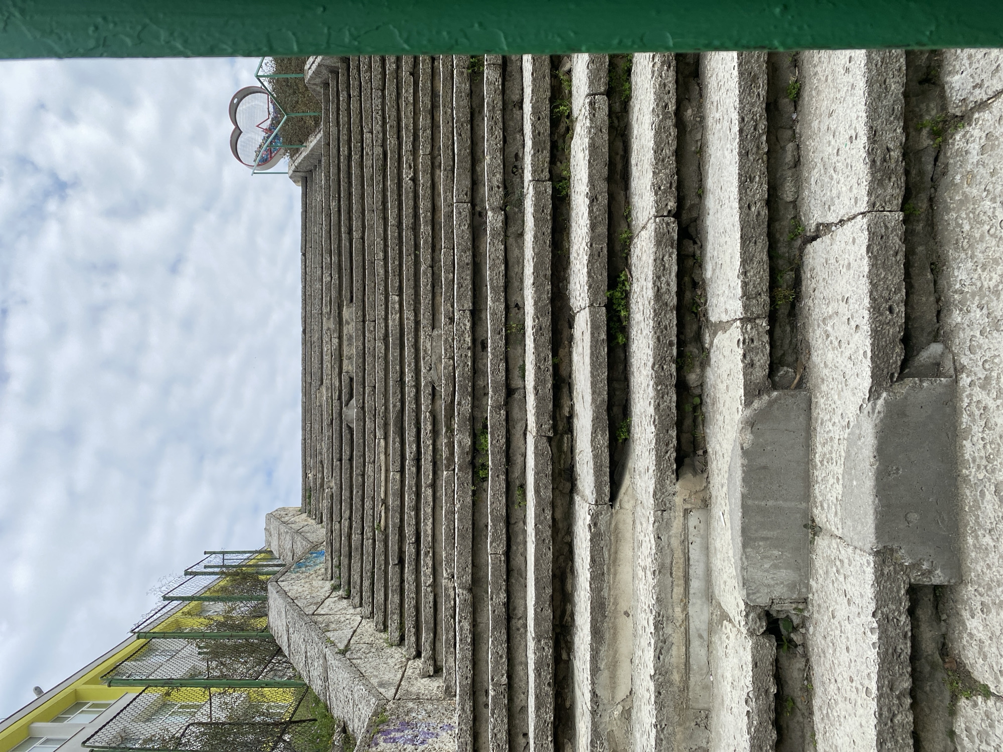 The image shows a long set of concrete stairs ascending upwards, with the view oriented looking up the steps. The steps are worn and show signs of age and weathering, with cracks and small patches of vegetation growing in between the steps. At the top of the stairs, a heart-shaped metal structure is visible. At the bottom of the stairs, a yellow and white building with a wire fence is partially visible. The sky is overcast with clouds. The steps are the Monument of the Bulgarian-Soviet Friendship in Varna, Bulgaria.