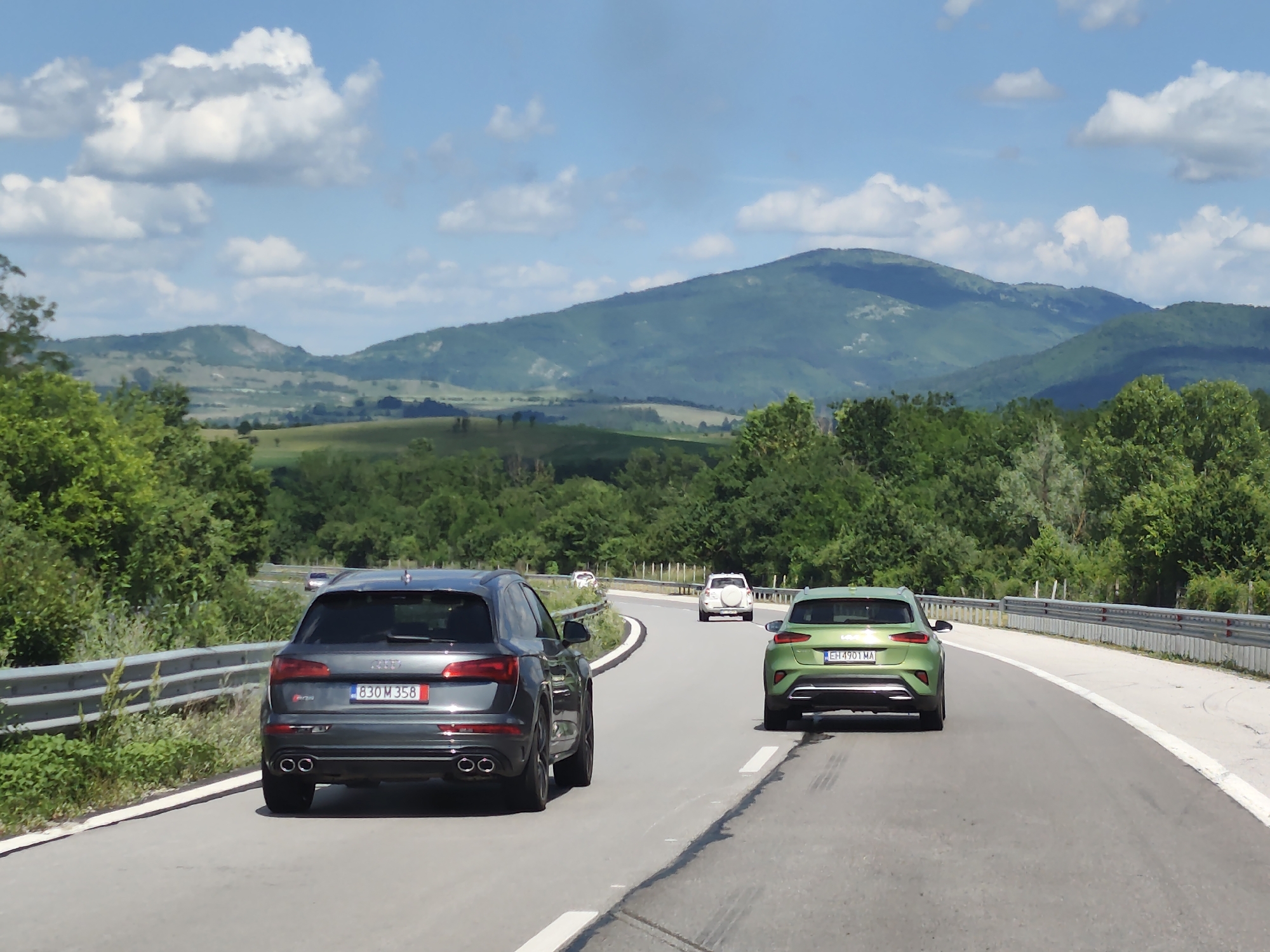 This media file depicts a scenic drive on a sunny day in Osikovitsa, Bulgaria. The scene is a paved road curving through rolling green hills and lush forests. In the foreground, two SUVs are driving away from the camera. The one on the left is a dark grey Audi, and the one on the right is a bright green Kia. Both vehicles are captured from the rear, showcasing their taillights and license plates. The Audi's license plate reads "830 M 358". The Kia's license plate is "EH 4901 MA".
Further down the road, other vehicles are visible, including a white SUV and a dark car in the distance. The sky is a vibrant blue, dotted with fluffy white clouds, suggesting pleasant weather. The surrounding landscape is characterized by verdant vegetation, with trees lining the sides of the road and hills stretching into the background. The time of day appears to be midday, given the bright sunlight casting shadows. The overall mood is one of leisurely travel and appreciation for the natural beauty of the Bulgarian countryside.